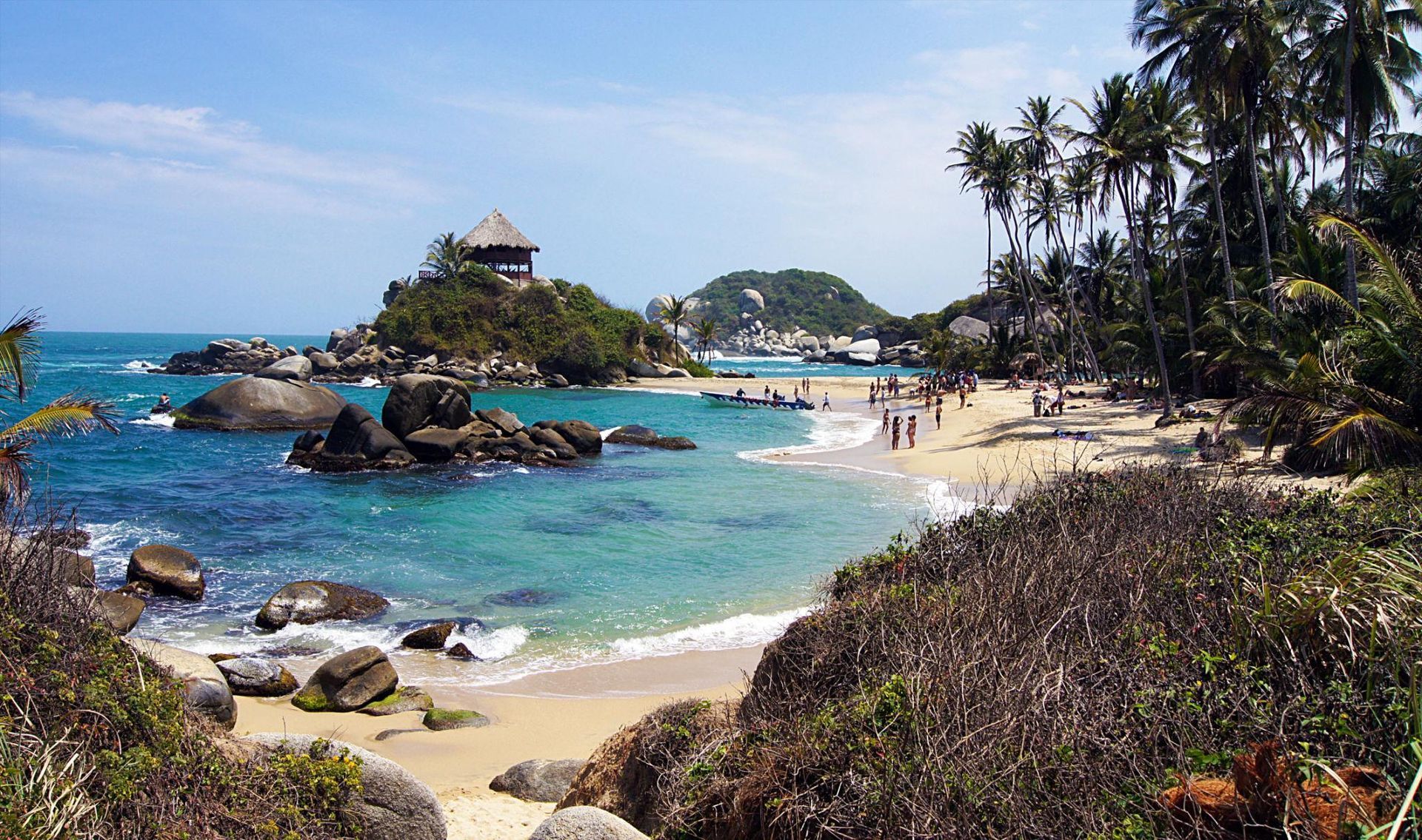 Tropical Caribbean beach with turquoise water, rocky shore, palm trees, and a small pavilion on a distant hill