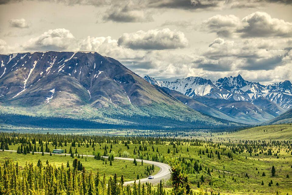 Winding road through green valley beneath snow-dusted mountains and cloudy sky