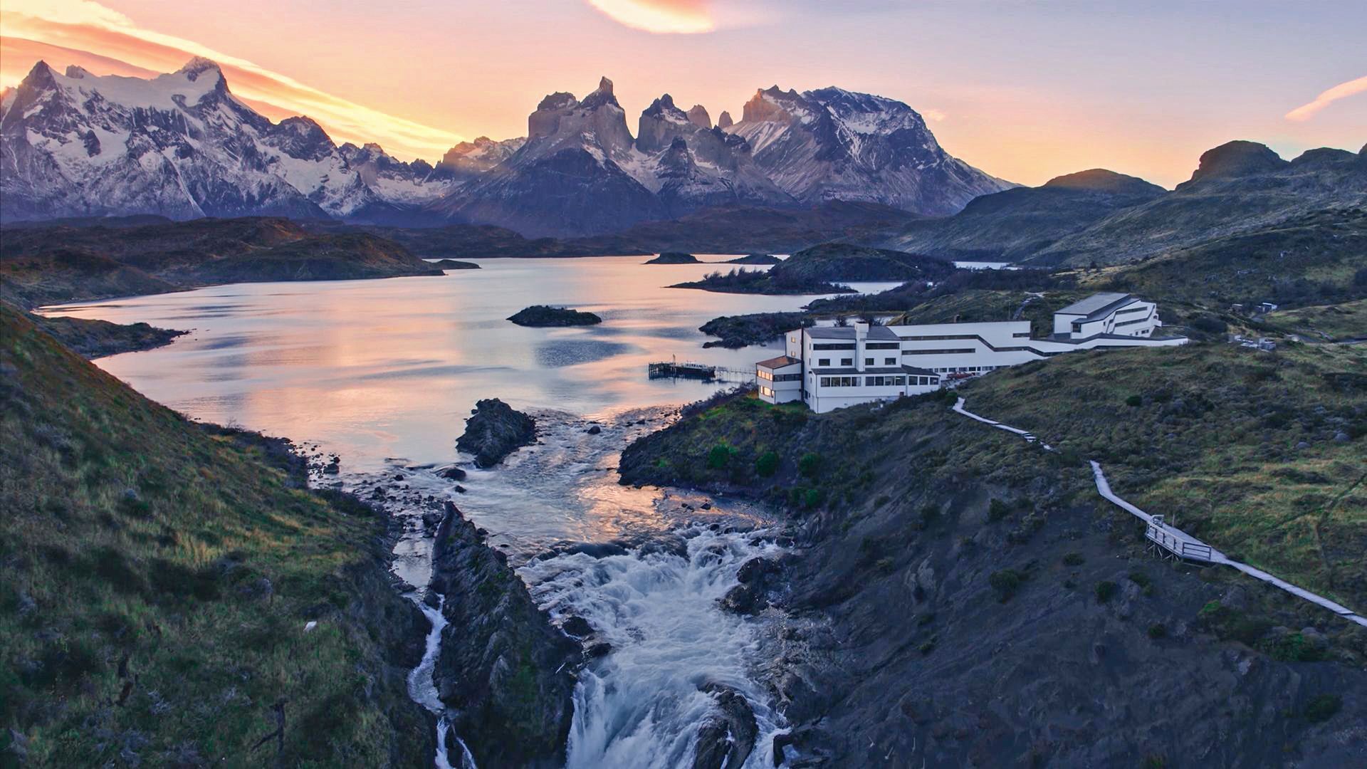 Mountain lake at sunset with snow-capped peaks, rocky cliffs, and buildings along the shore