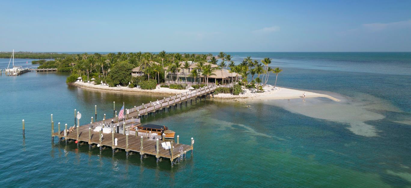 Aerial view of a tropical island resort with a long pier, palm trees, and turquoise water