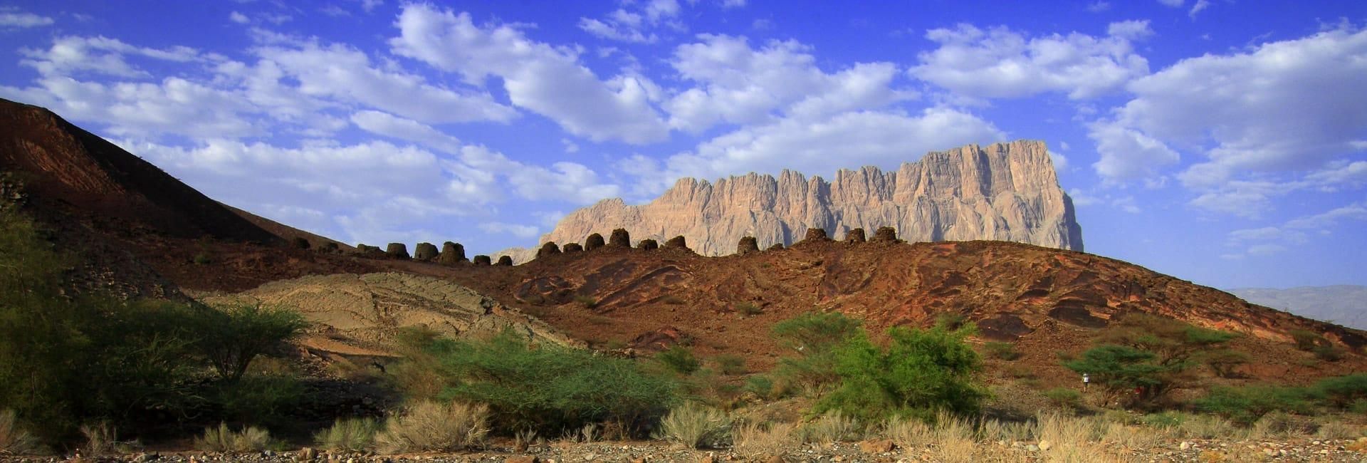 Rocky desert hills under a blue sky with clouds and a line of rugged cliffs in the distance
