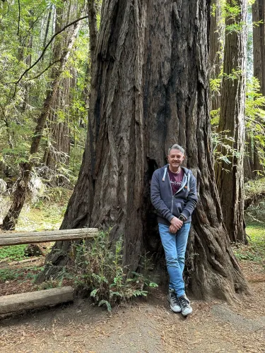 Man leans against a large redwood tree in a forest. He wears a jacket, jeans, and sneakers.