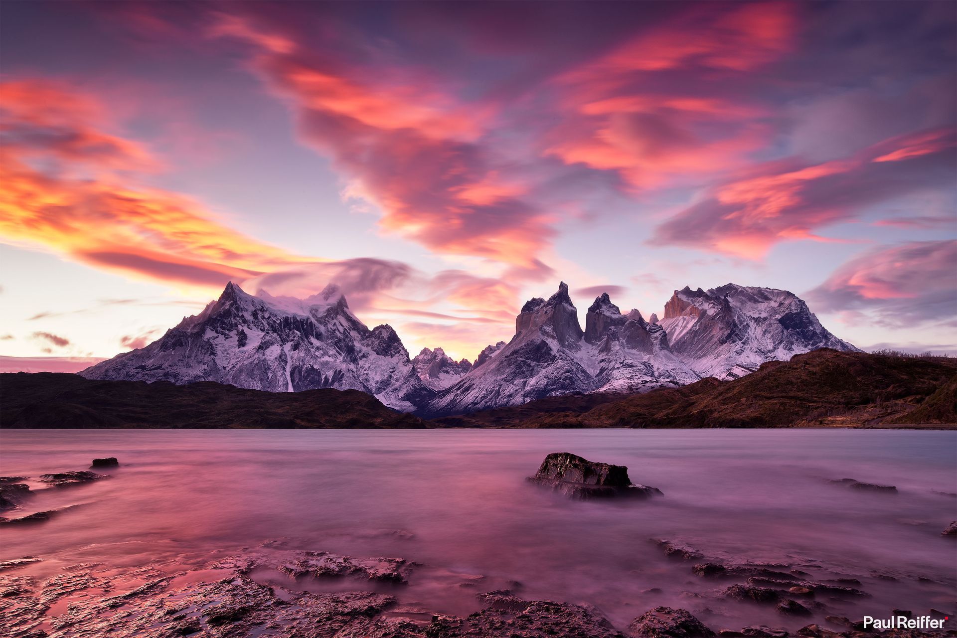 Snowy mountain peaks at sunset above a pink reflective lake under dramatic clouds.