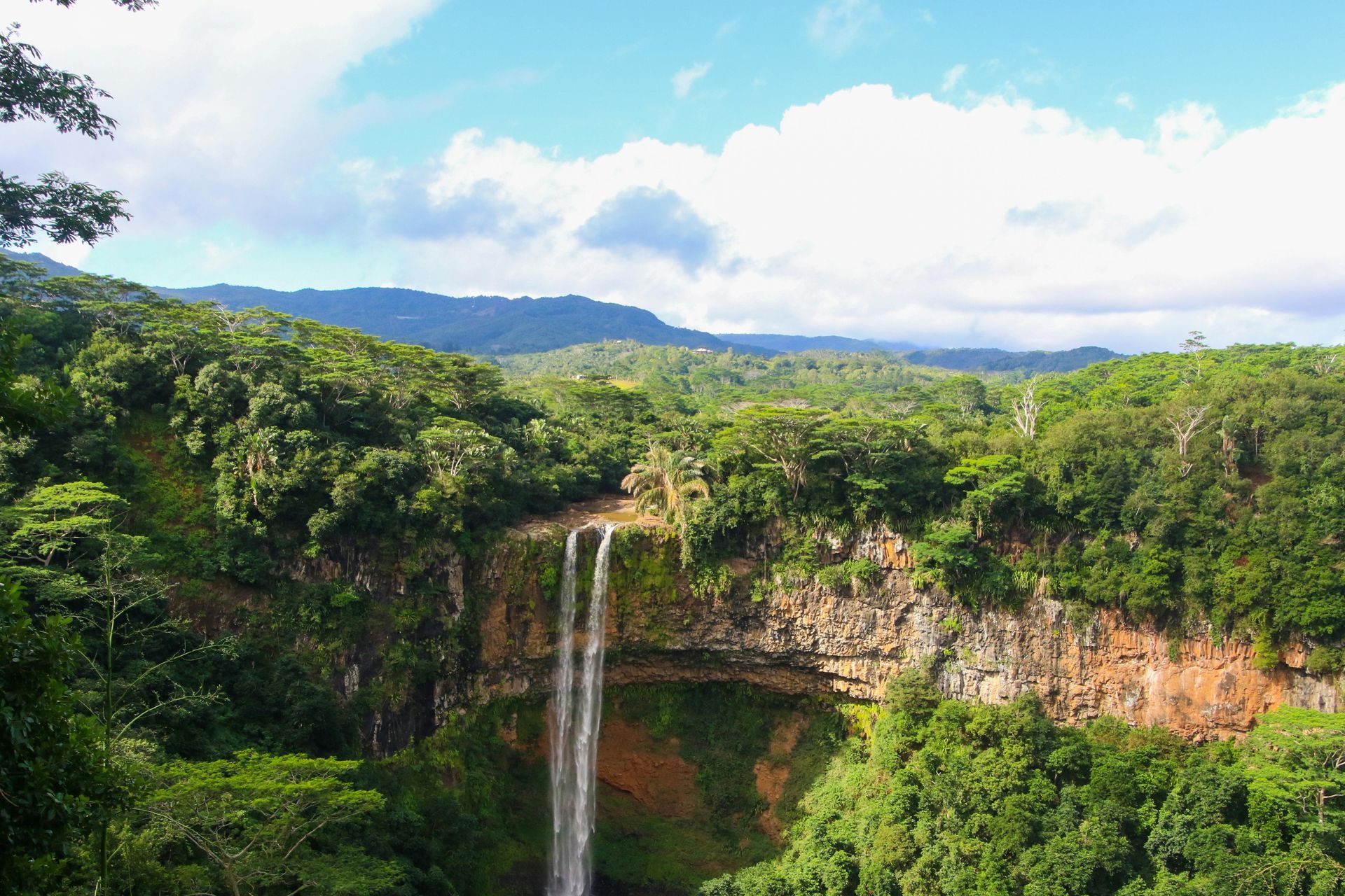 Waterfall cascading down a rocky cliff, surrounded by lush green foliage and mountains under a blue sky.