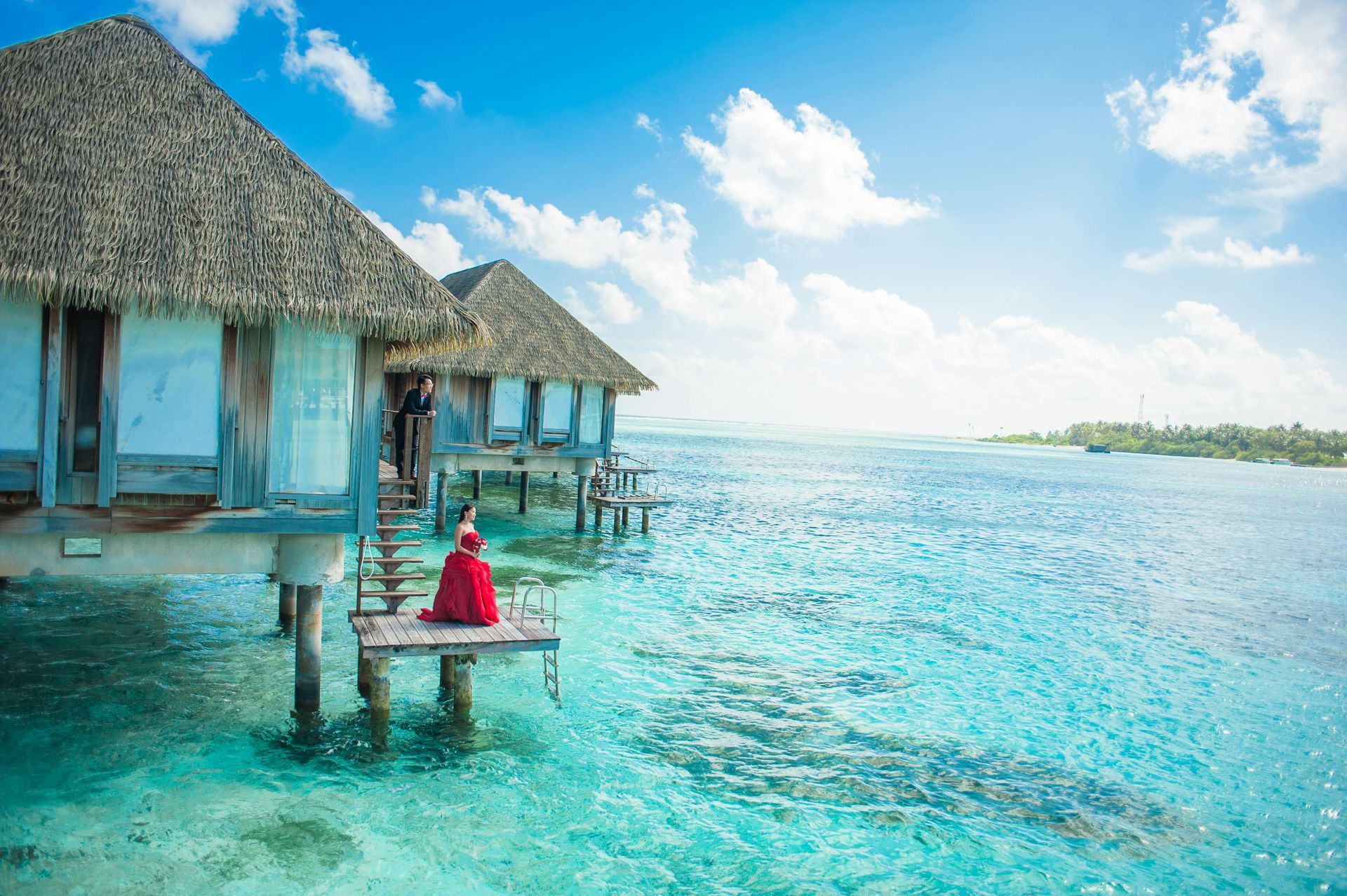 Overwater bungalows on stilts in clear turquoise water; person in red sits on a deck, sunny sky.