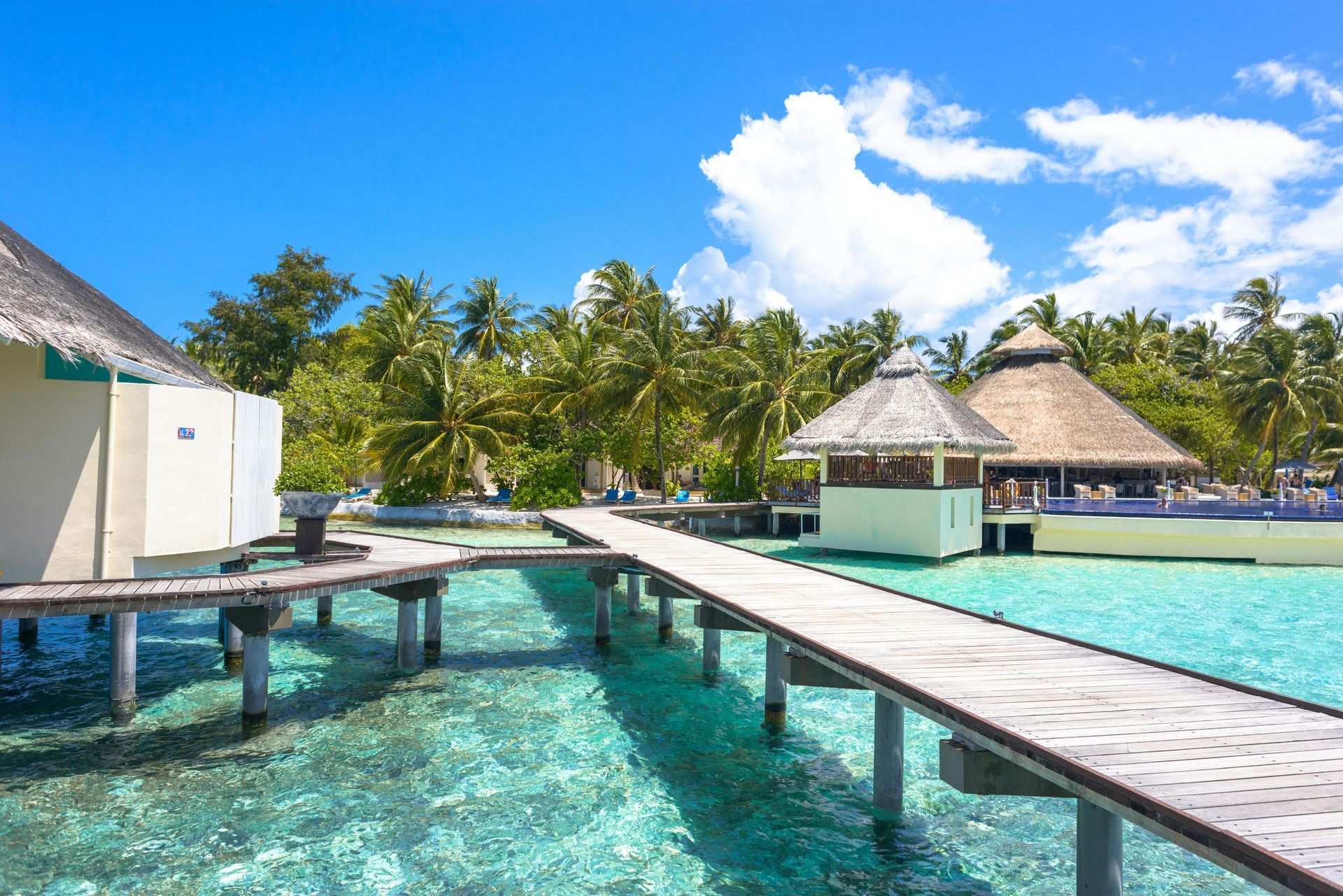 Wooden walkways over turquoise water, leading to overwater bungalows and a thatched-roof structure under a bright blue sky.