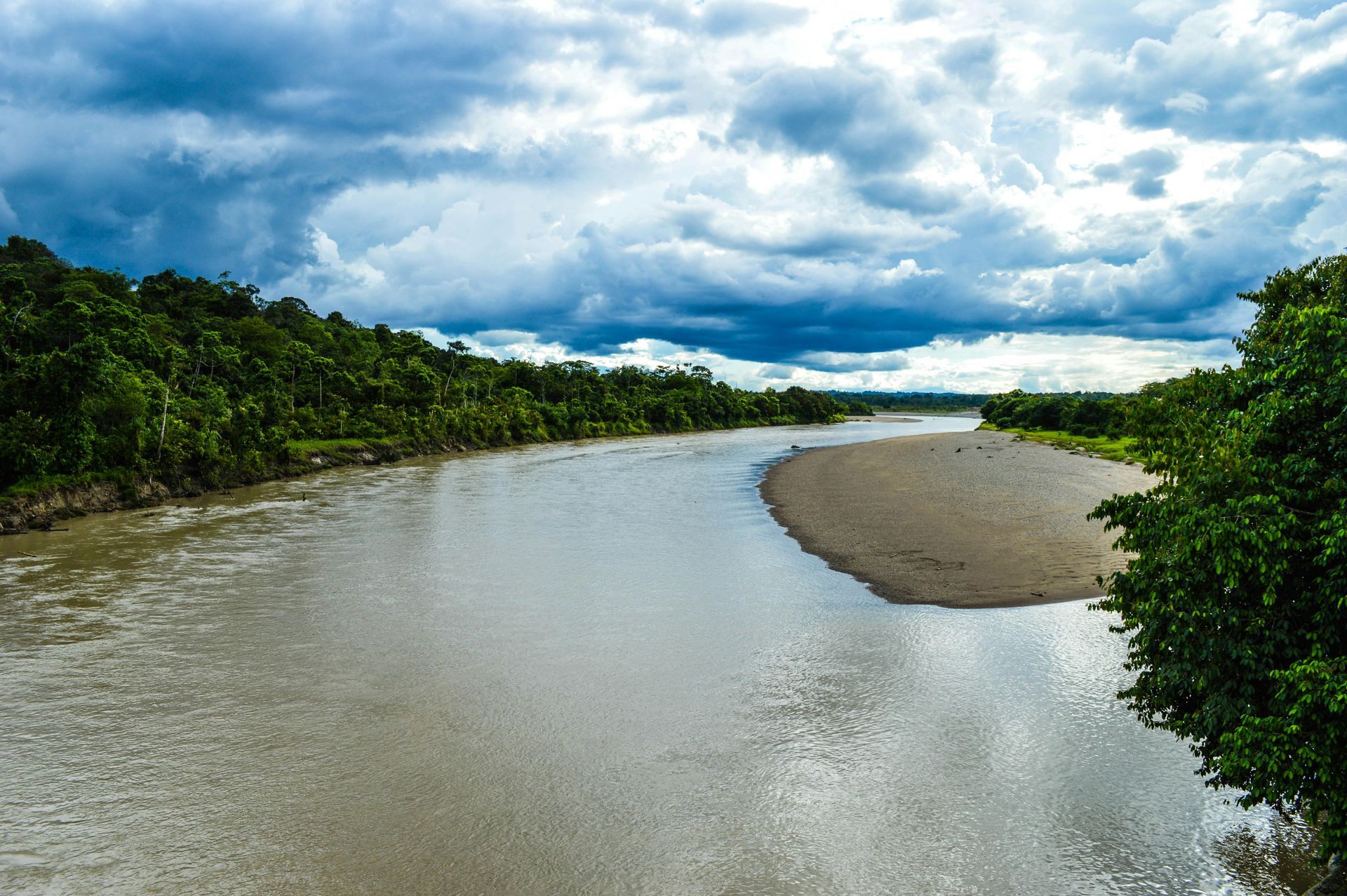 Wide river winding through lush green forest under a cloudy sky, with a sandy bank on the right