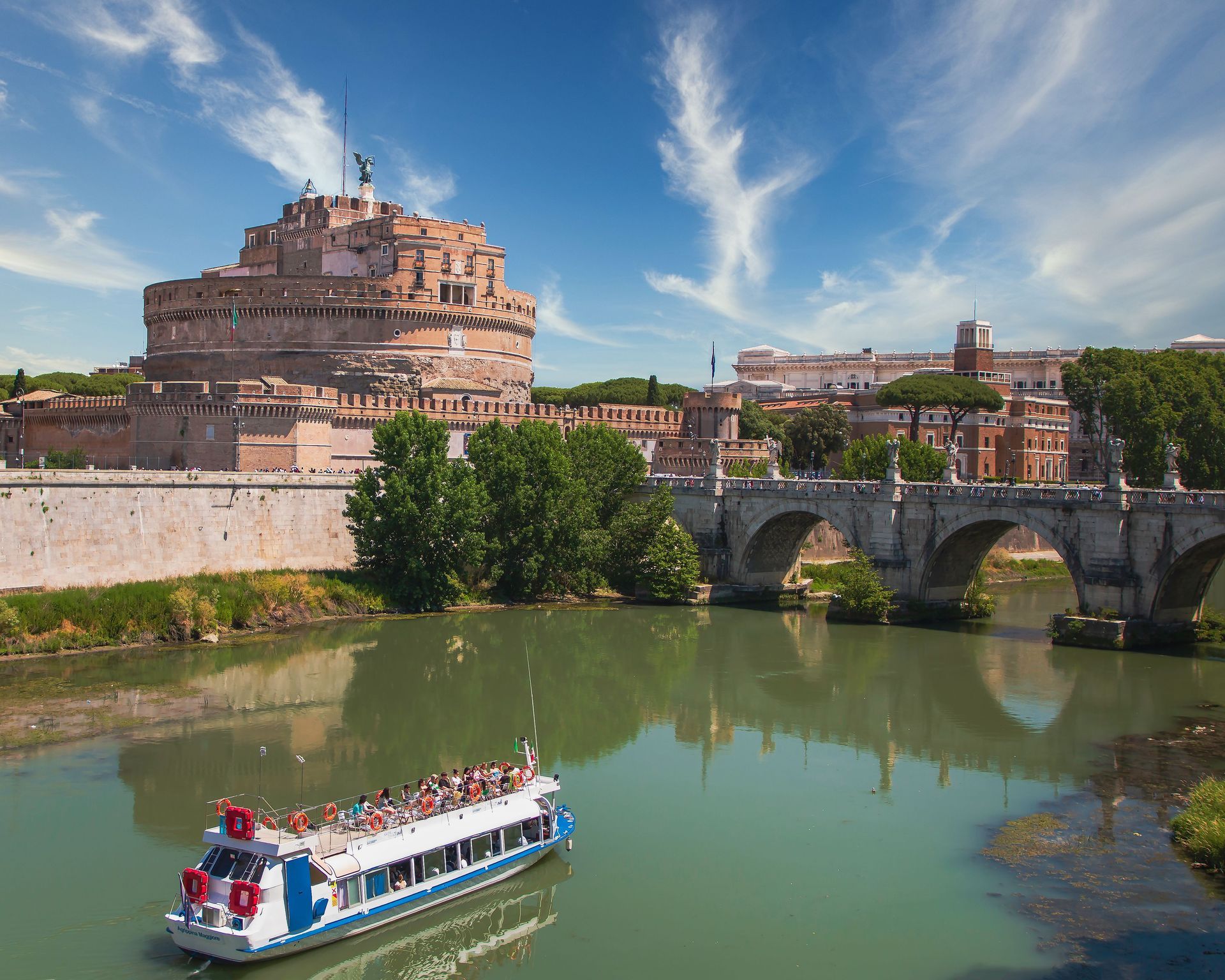 Riverboat on green river beside a stone bridge and historic fortress under a blue sky