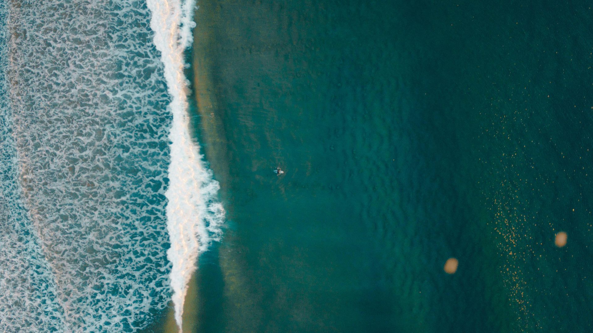 Overhead view: Ocean waves breaking on the shore, with a distant surfer in the teal water.