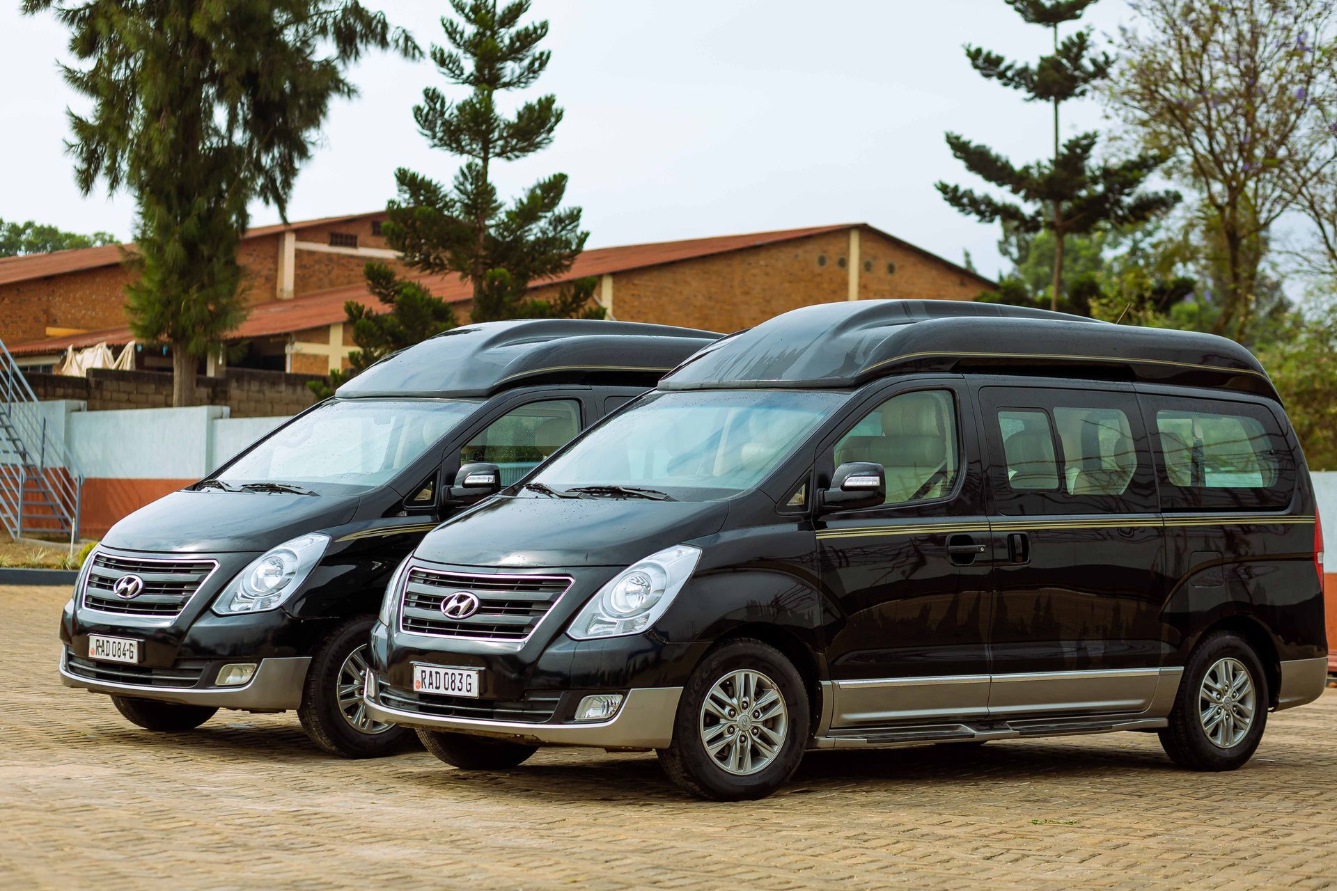 Two black vans parked outdoors in front of a brick building and trees