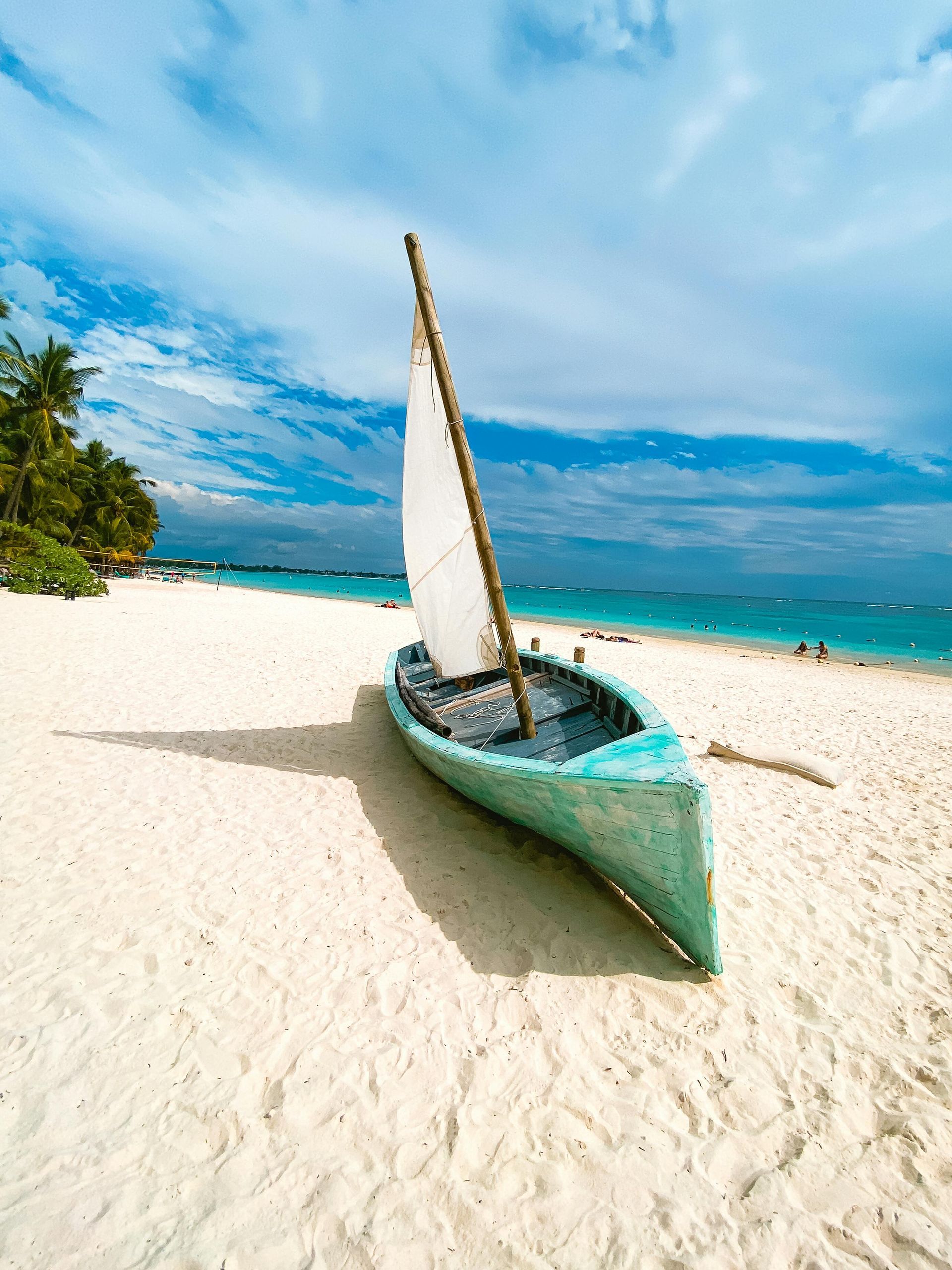 Light blue boat with sail on a white sandy beach, under a blue sky.