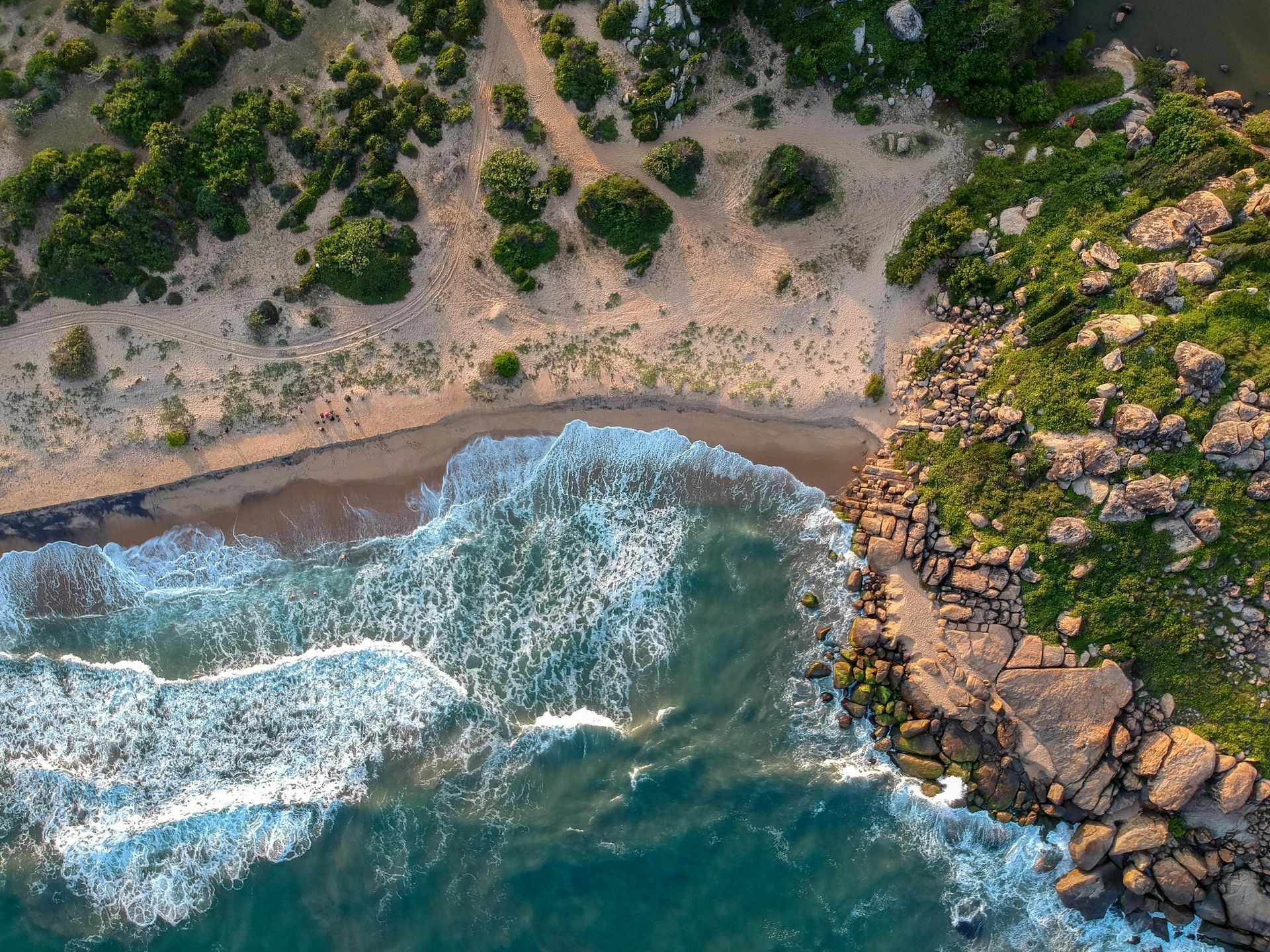 Aerial view of a sandy beach, rocky coastline, and ocean waves. Green foliage lines the shore.