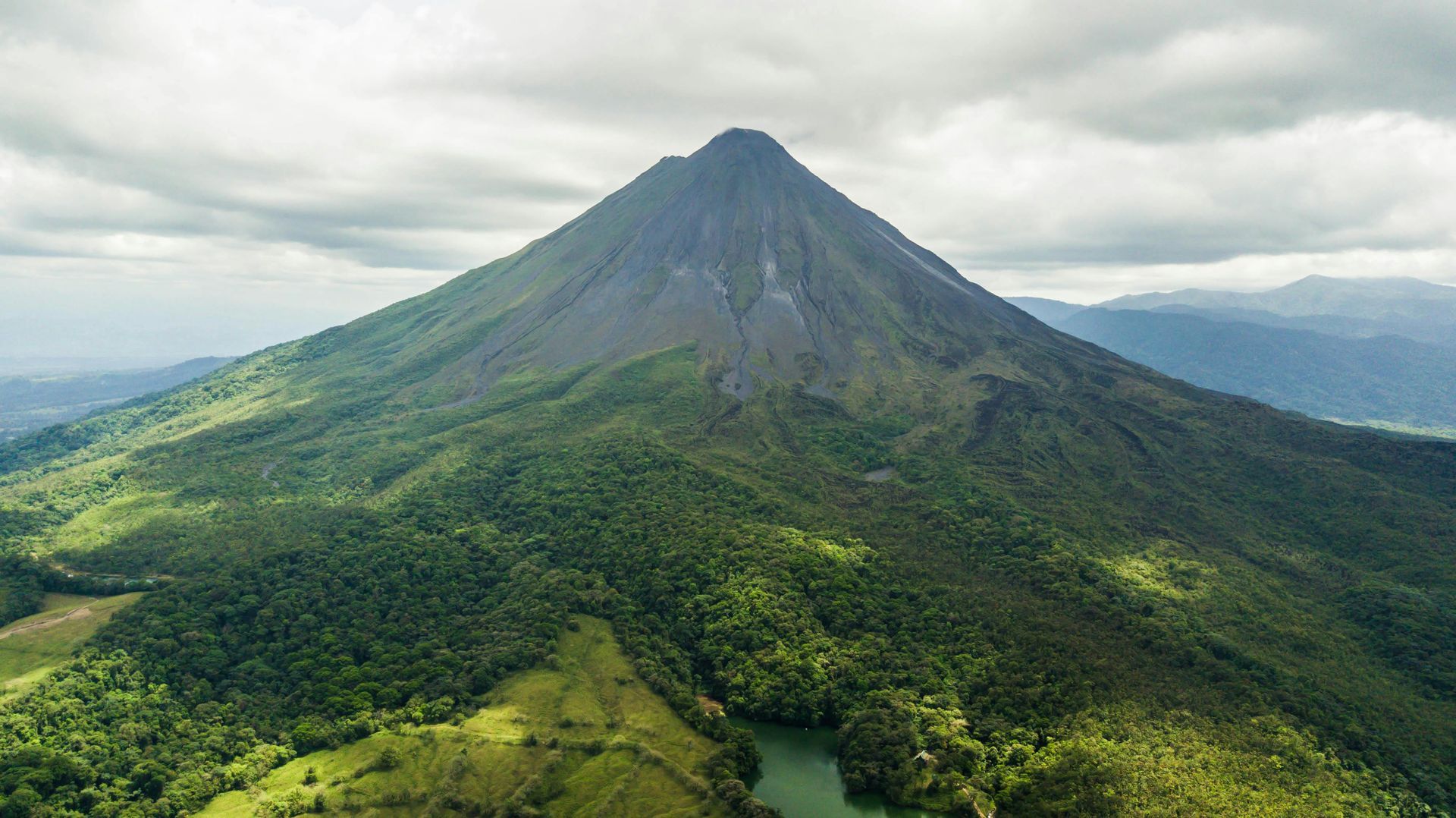 A lush green volcano with a cone shape rises under cloudy skies.