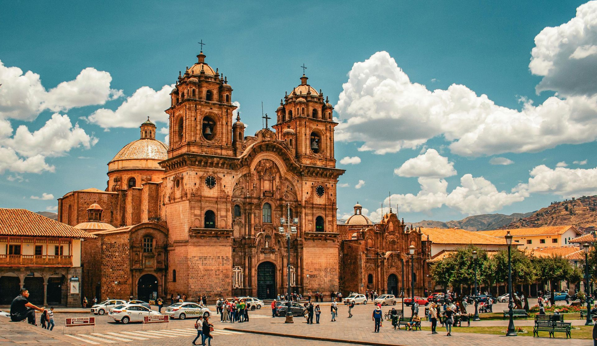 Historic cathedral with twin domes in a sunny plaza, people walking below and mountains in the background