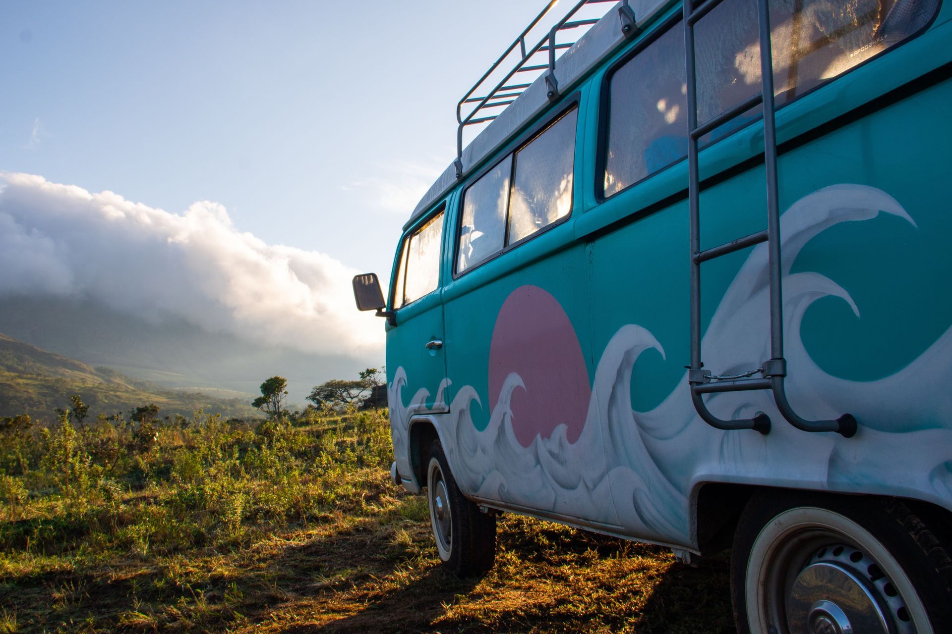 Teal van with wave mural parked beside a grassy hillside at sunset