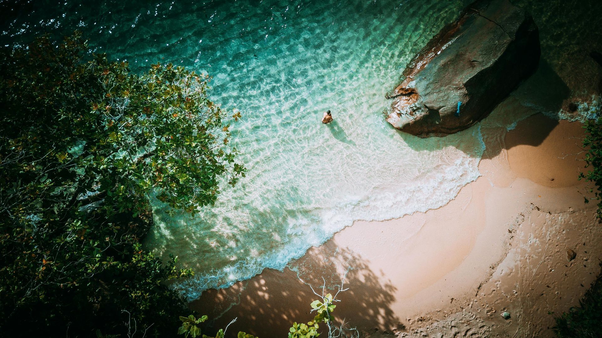 Person wading in clear turquoise water near a sandy beach and large rock; overhead shot.