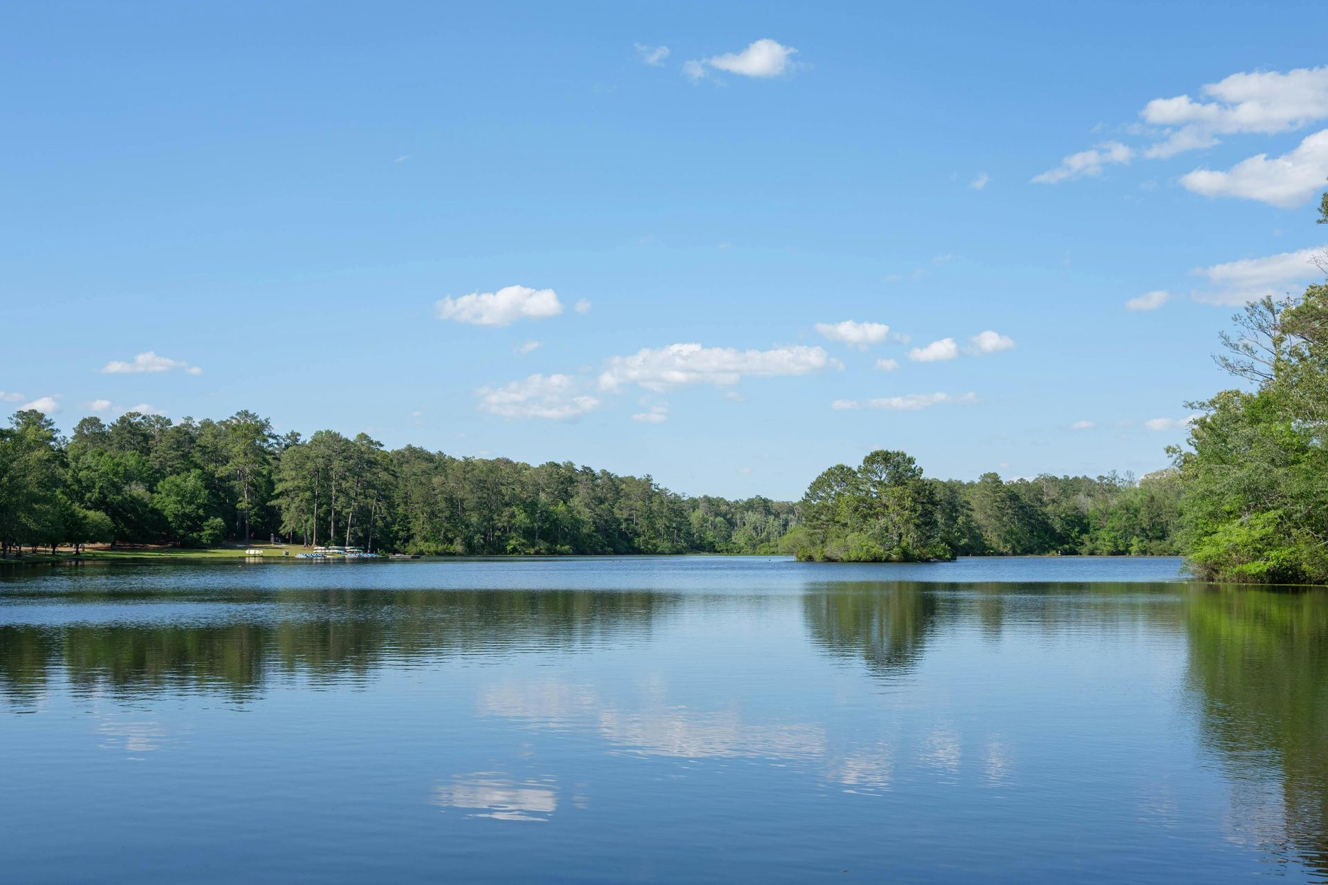 Calm lake reflecting a blue sky with scattered clouds, surrounded by green trees.