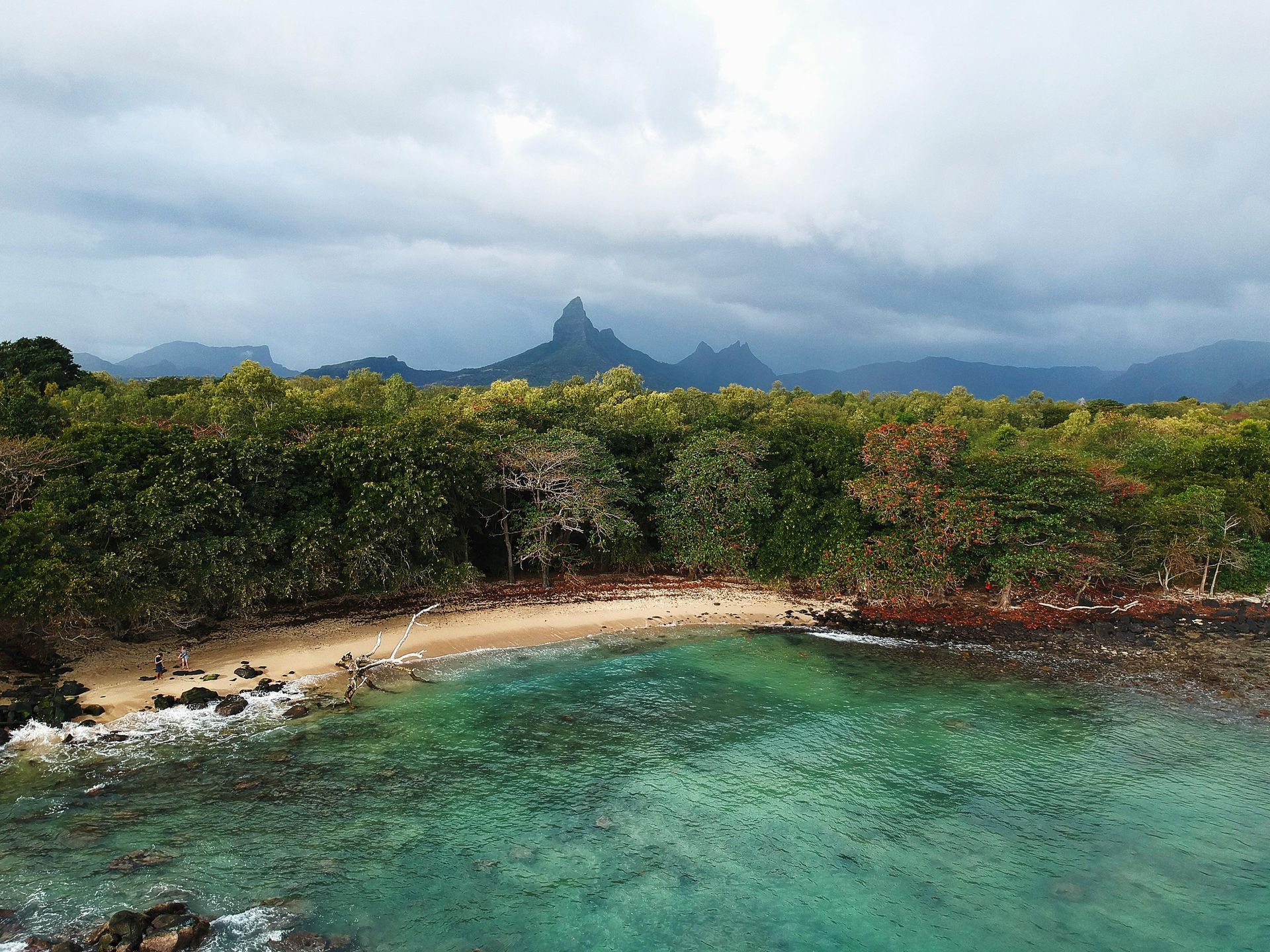 Tropical beach with turquoise water, sandy shore, green trees, and mountains under cloudy sky.