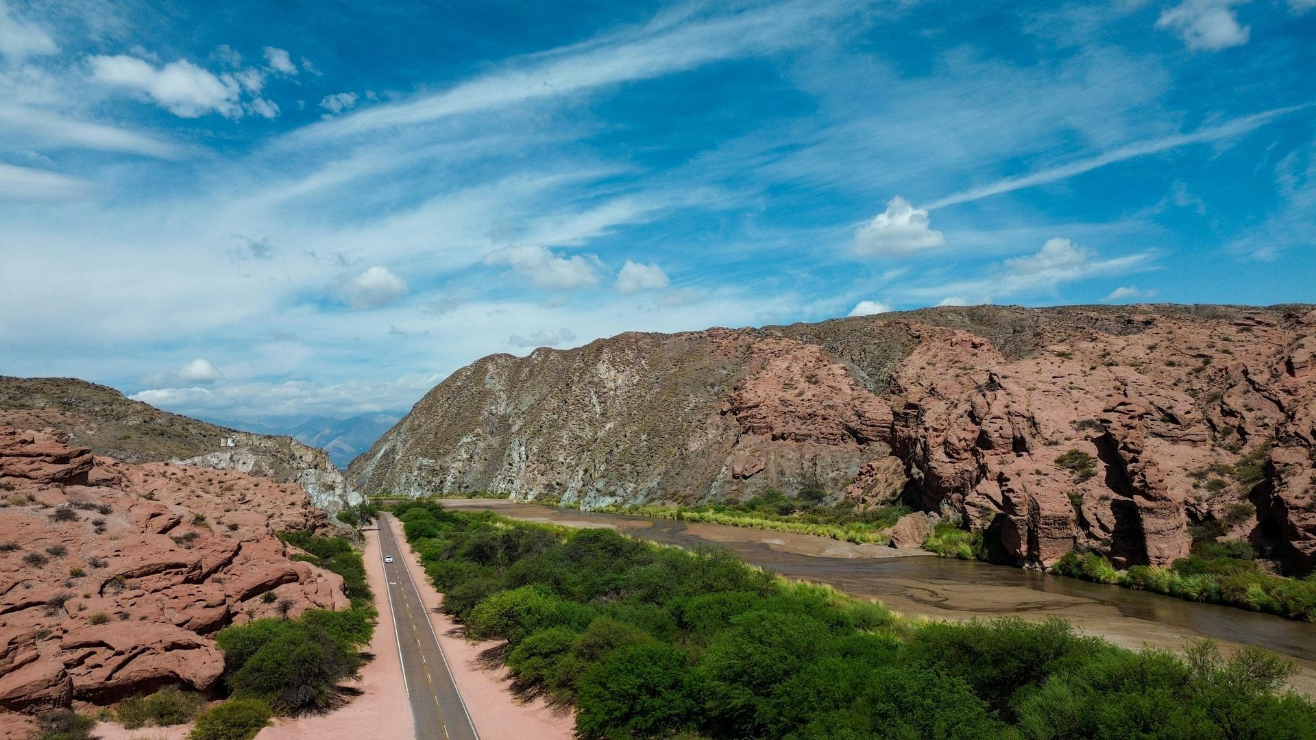 Road winds through a canyon, river and green vegetation below. Red rock cliffs and a blue sky above.