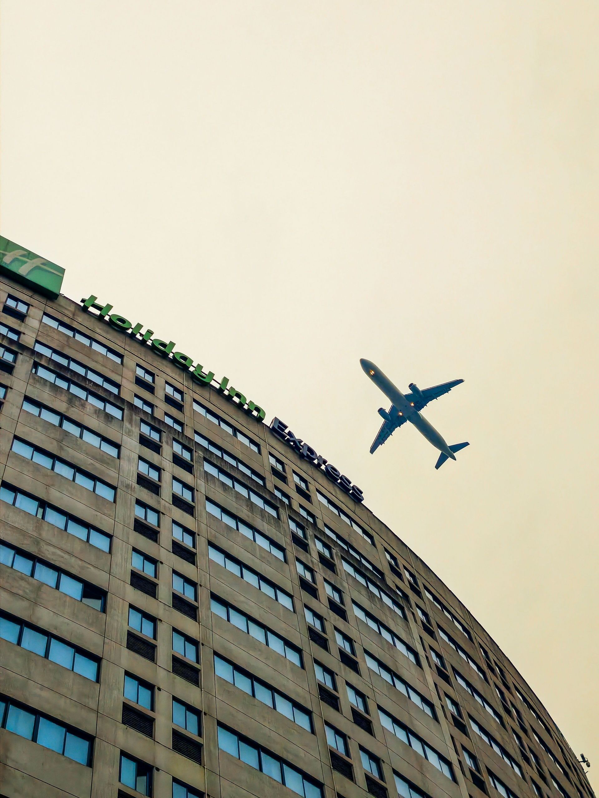 Airplane flying above a curved high-rise building against a pale sky