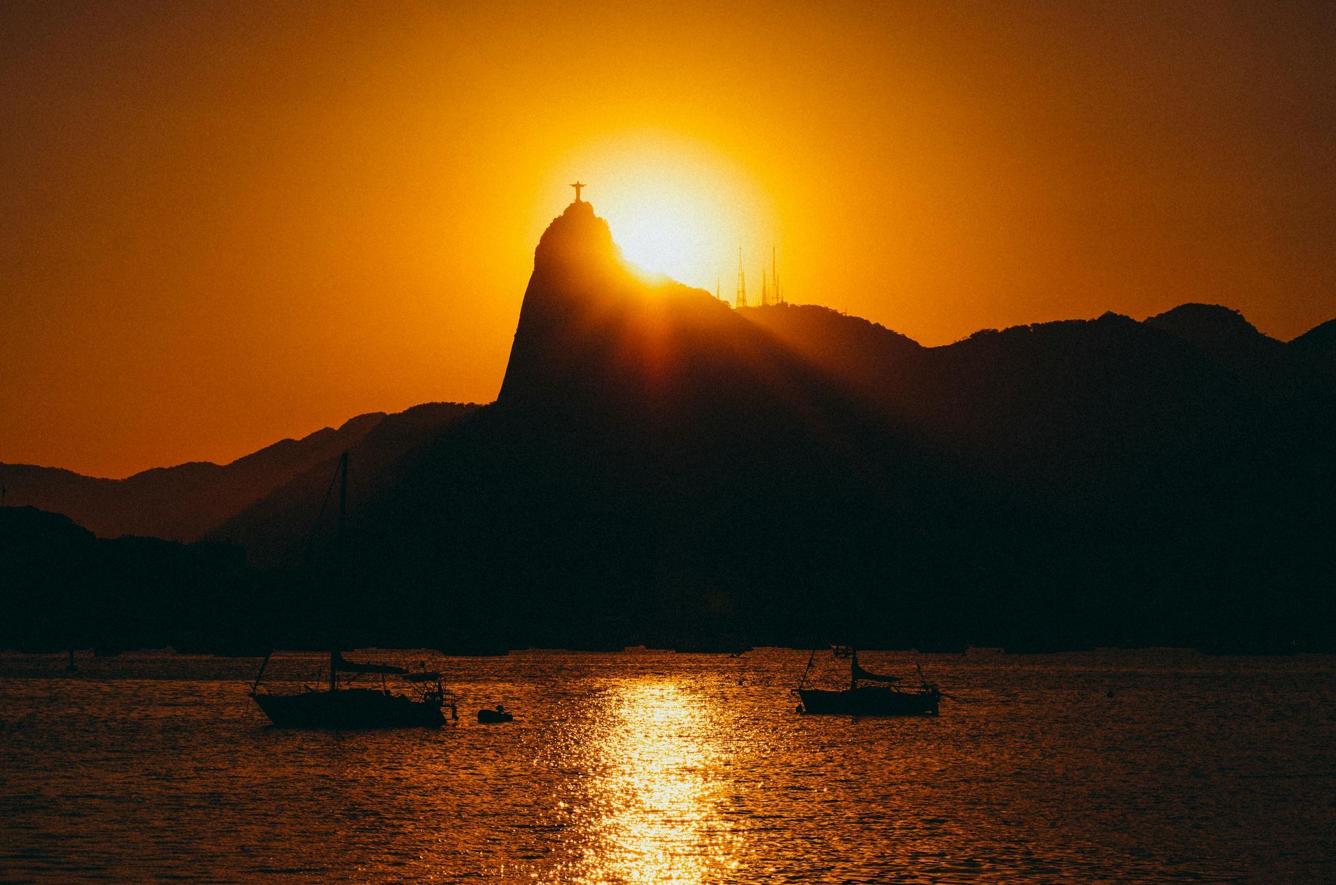 Sunset over Rio de Janeiro, silhouette of Christ the Redeemer statue on Corcovado Mountain, boats on water.