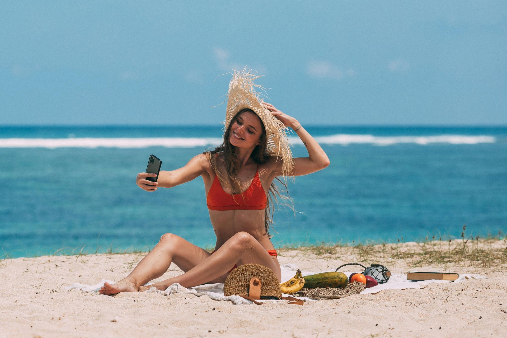 Woman in a straw hat sitting on a beach, smiling and holding a drink with ocean behind her.