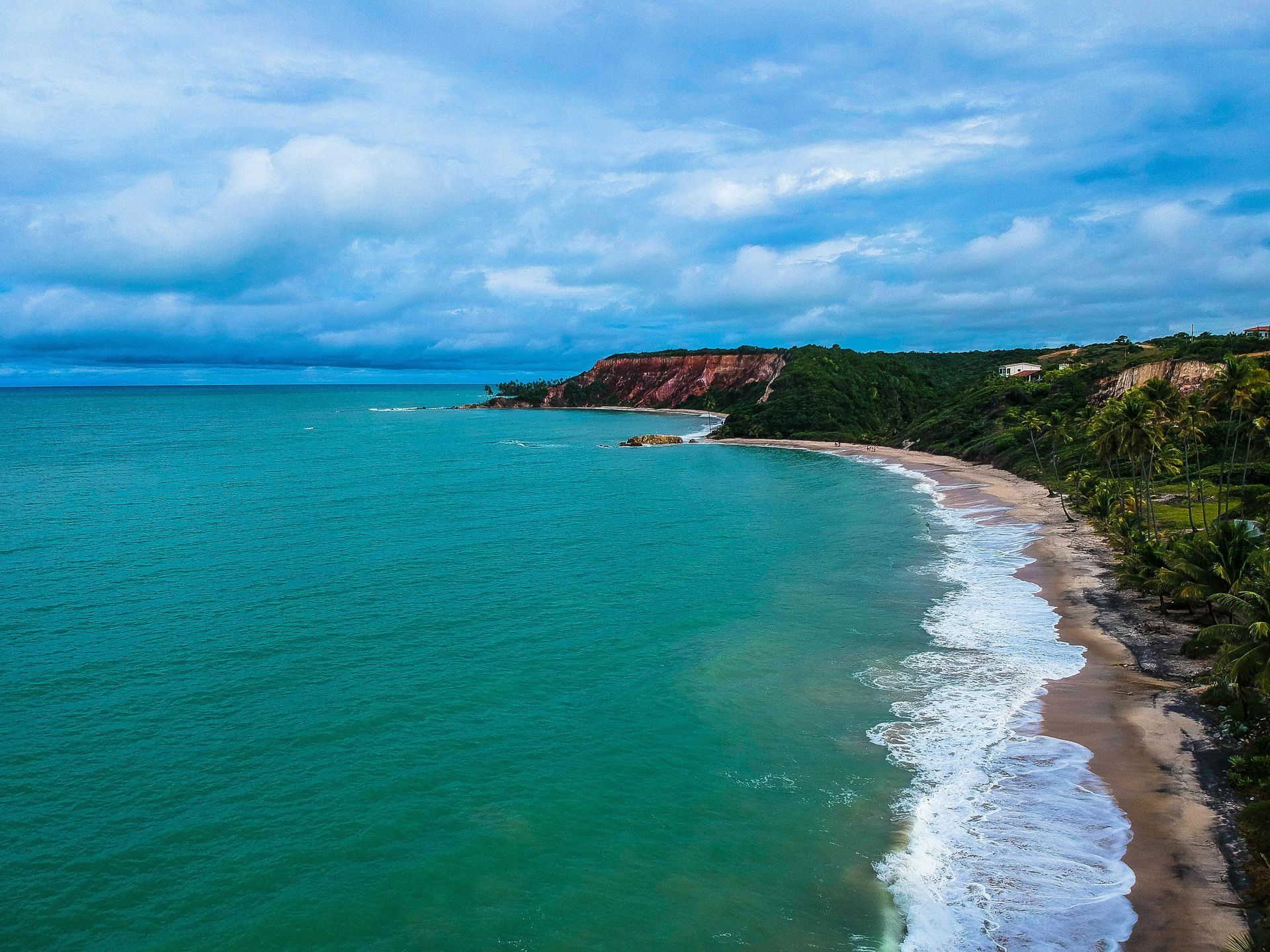 Turquoise ocean waves crashing on a sandy beach, with a lush green cliffside under a cloudy sky.