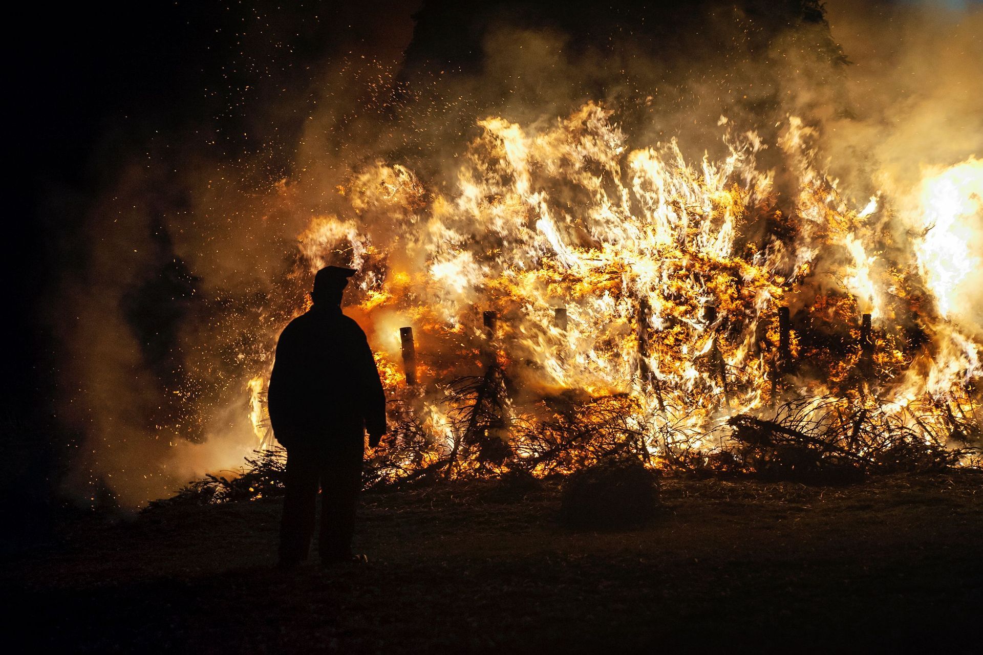 Silhouetted person facing a large bonfire with bright flames and smoke at night