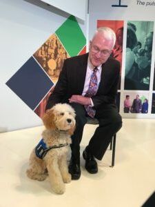 A smiling person in a dark suit and patterned tie sitting in a chair, patting a seated light-colored dog in a harness.