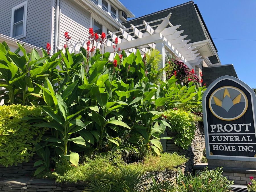 A sign for Prout Funeral Home Inc. next to a garden of green plants and red flowers in front of a white building.