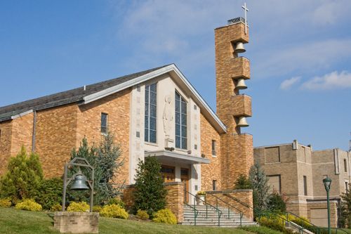 A tan brick church building featuring a bell tower with exposed bells, a stone staircase, and a decorative outdoor bell.