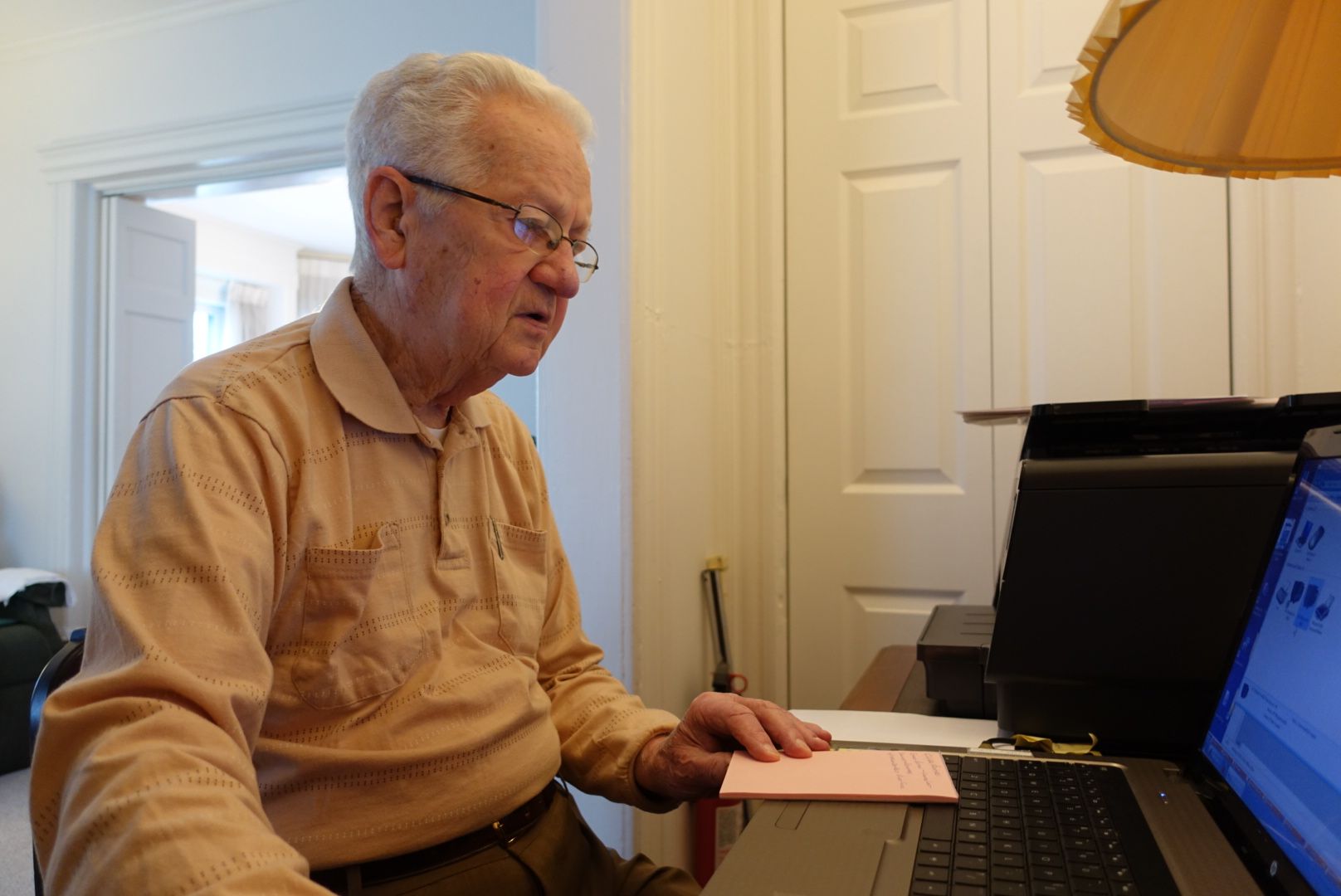 A person wearing glasses and a tan shirt sits at a desk, looking at a laptop computer next to a small pink notepad.