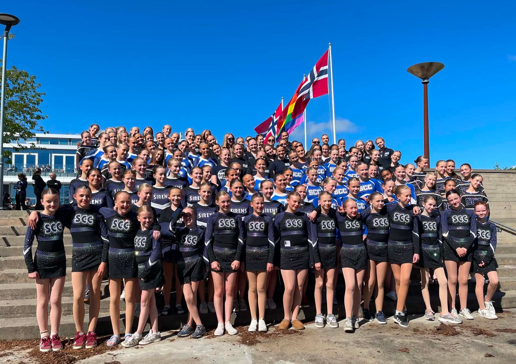 Large group in matching black shirts posing on outdoor steps, with a Norwegian flag and blue sky behind them