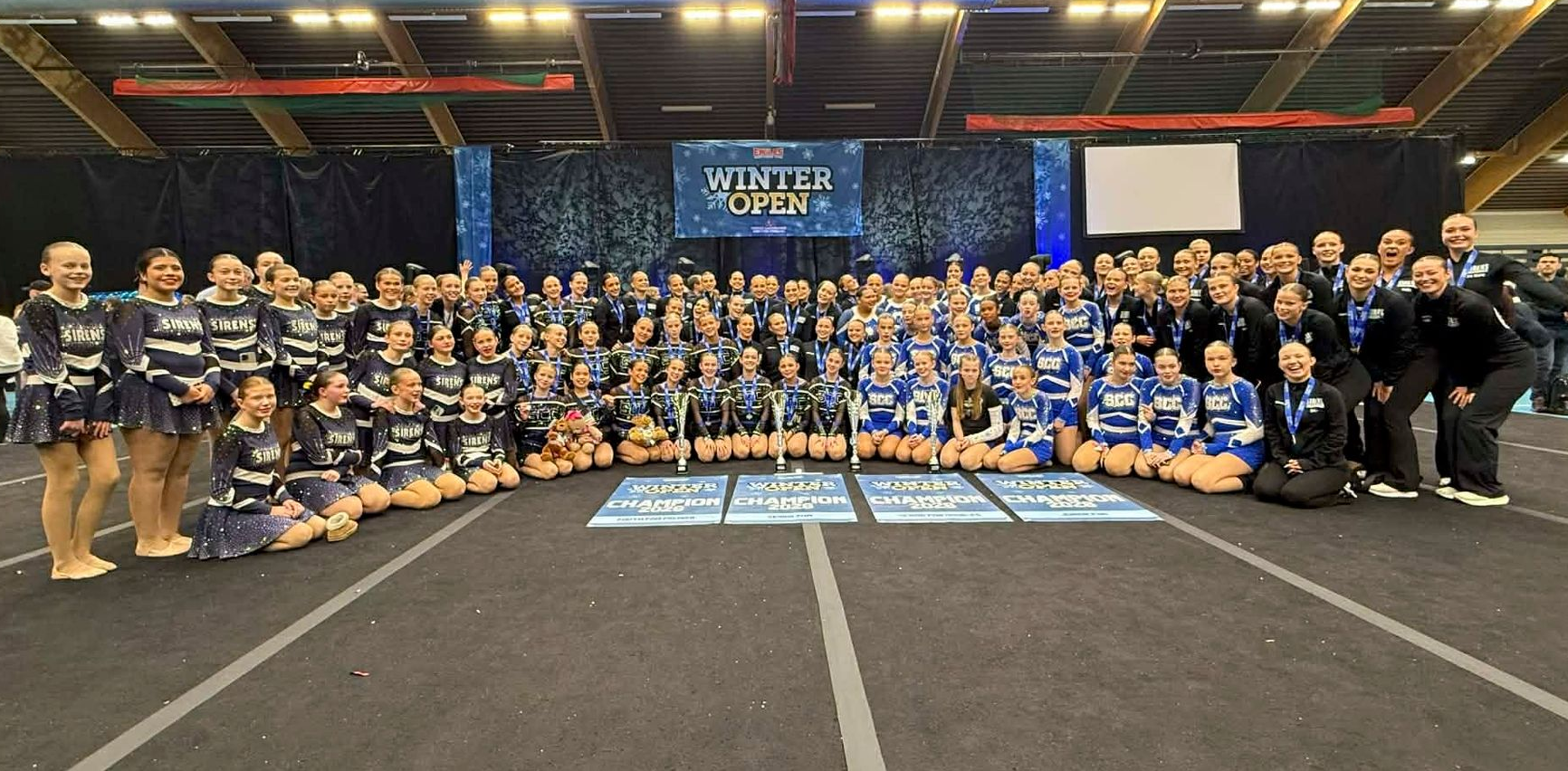Large group of cheerleaders in matching blue and white uniforms posing indoors with a banner.