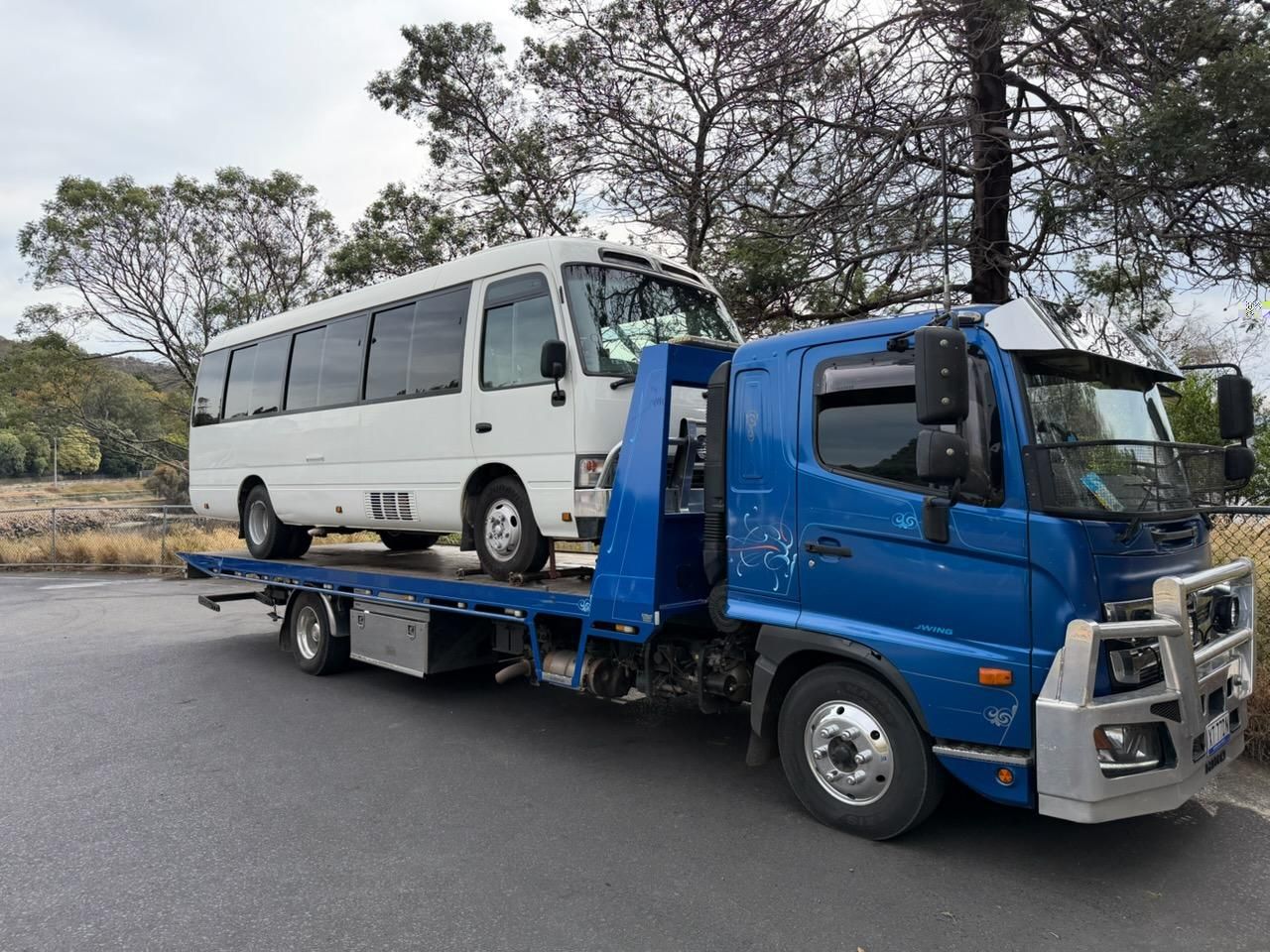 Blue flatbed tow truck carrying a white minibus on a roadside with trees nearby — AAA ULTRA TOWING in Mornington, TAS