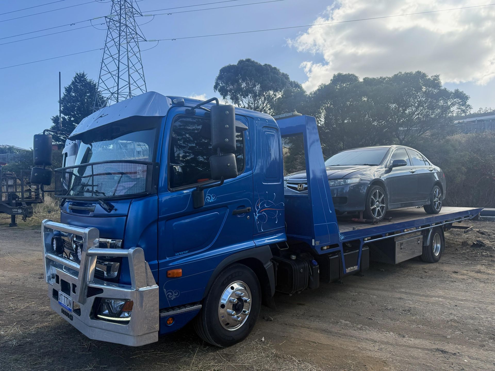 Blue car carrier truck loaded with a silver sedan on a dirt lot under a cloudy sky