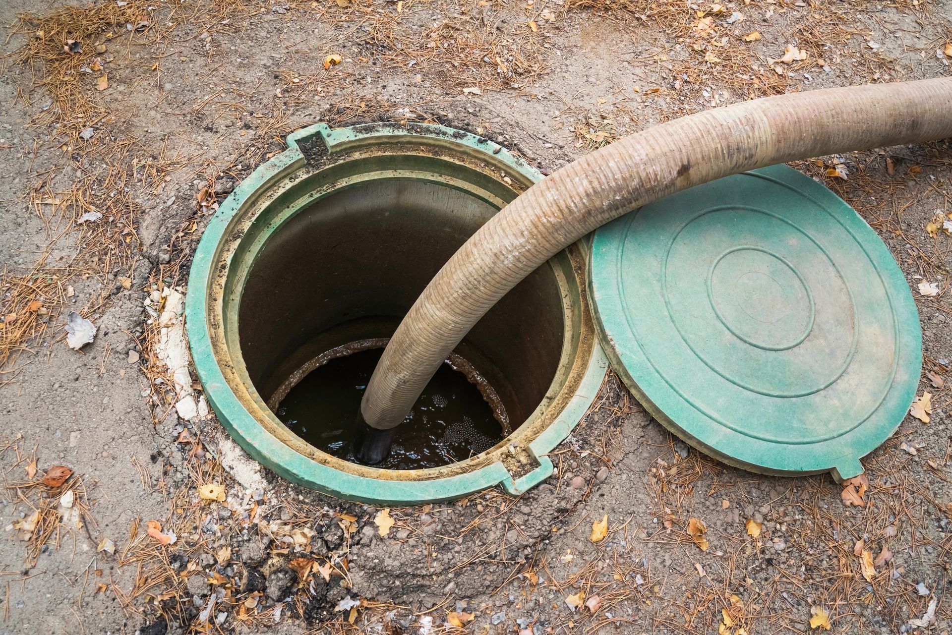 Open sewer manhole with a green cover and hose inserted into it on a dirt ground