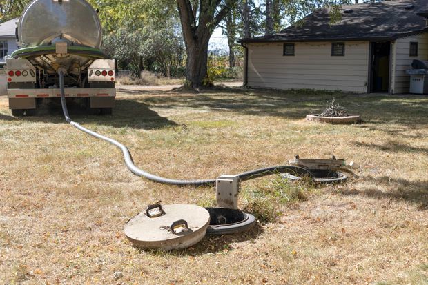 Yard with a low pipe or hose leading from a tank to two round concrete lids near a house and trees