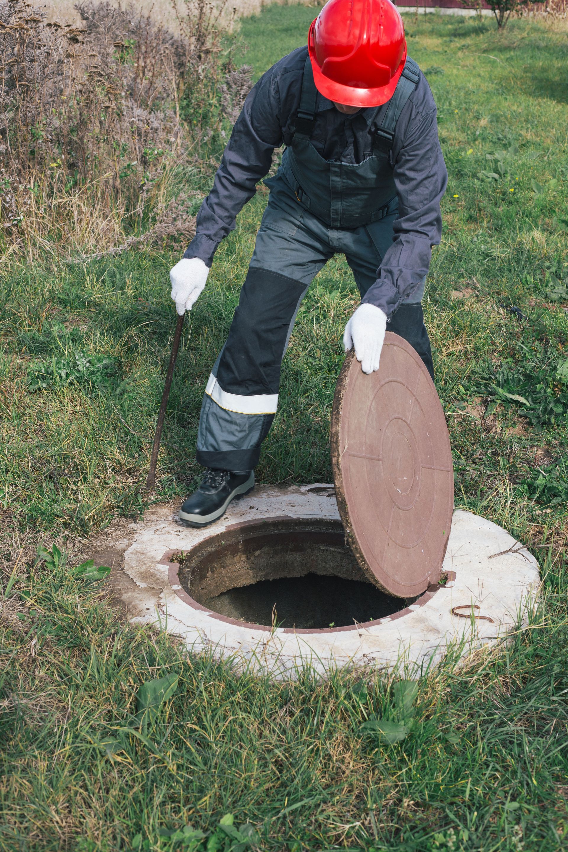 Worker in red hard hat opening a manhole cover on grassy ground