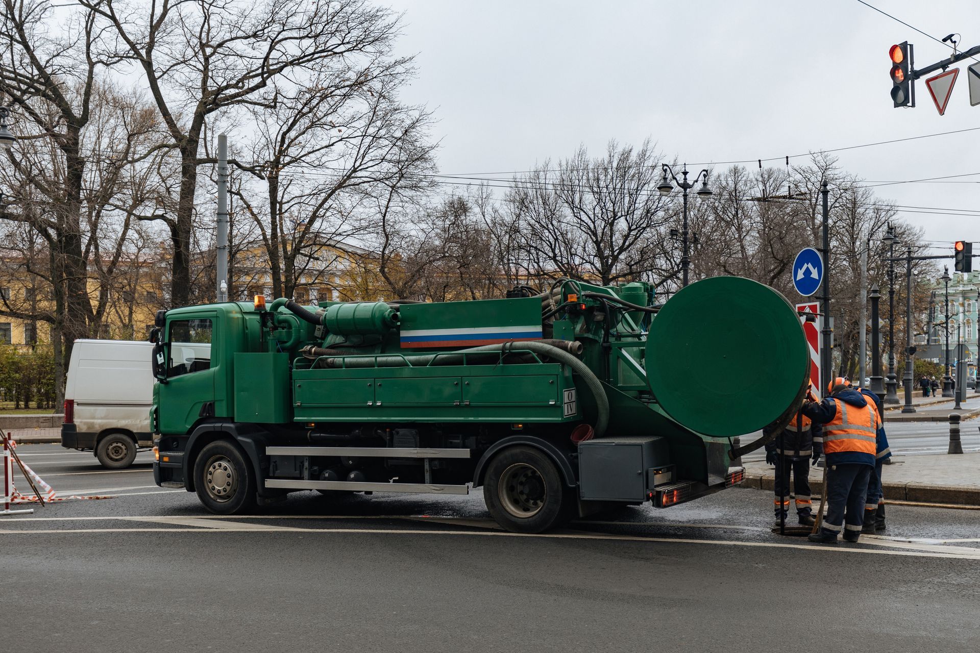 Green vacuum truck at a wet roadside with two workers near a traffic sign