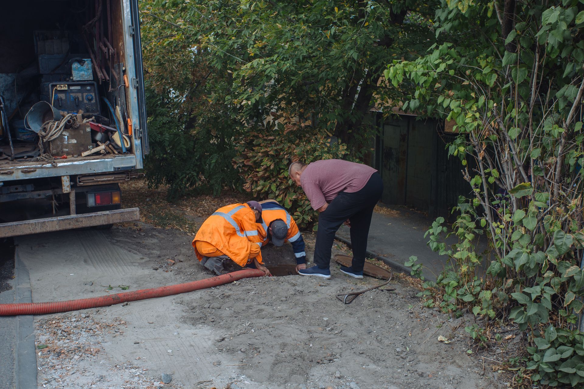 Two workers beside a truck use a hose on wet ground near dense greenery