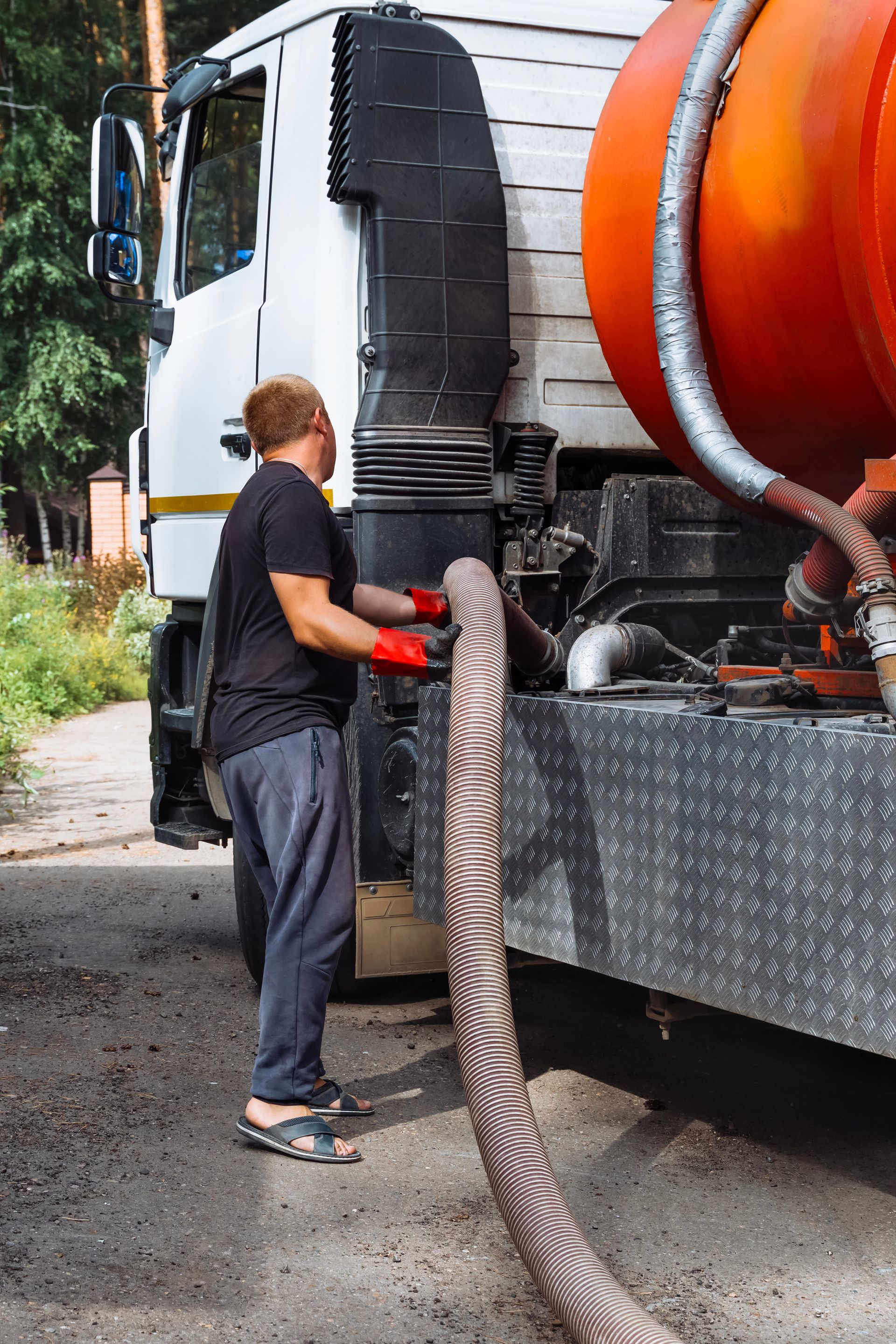 Worker handling a large hose beside a truck with an orange tank on a roadside.