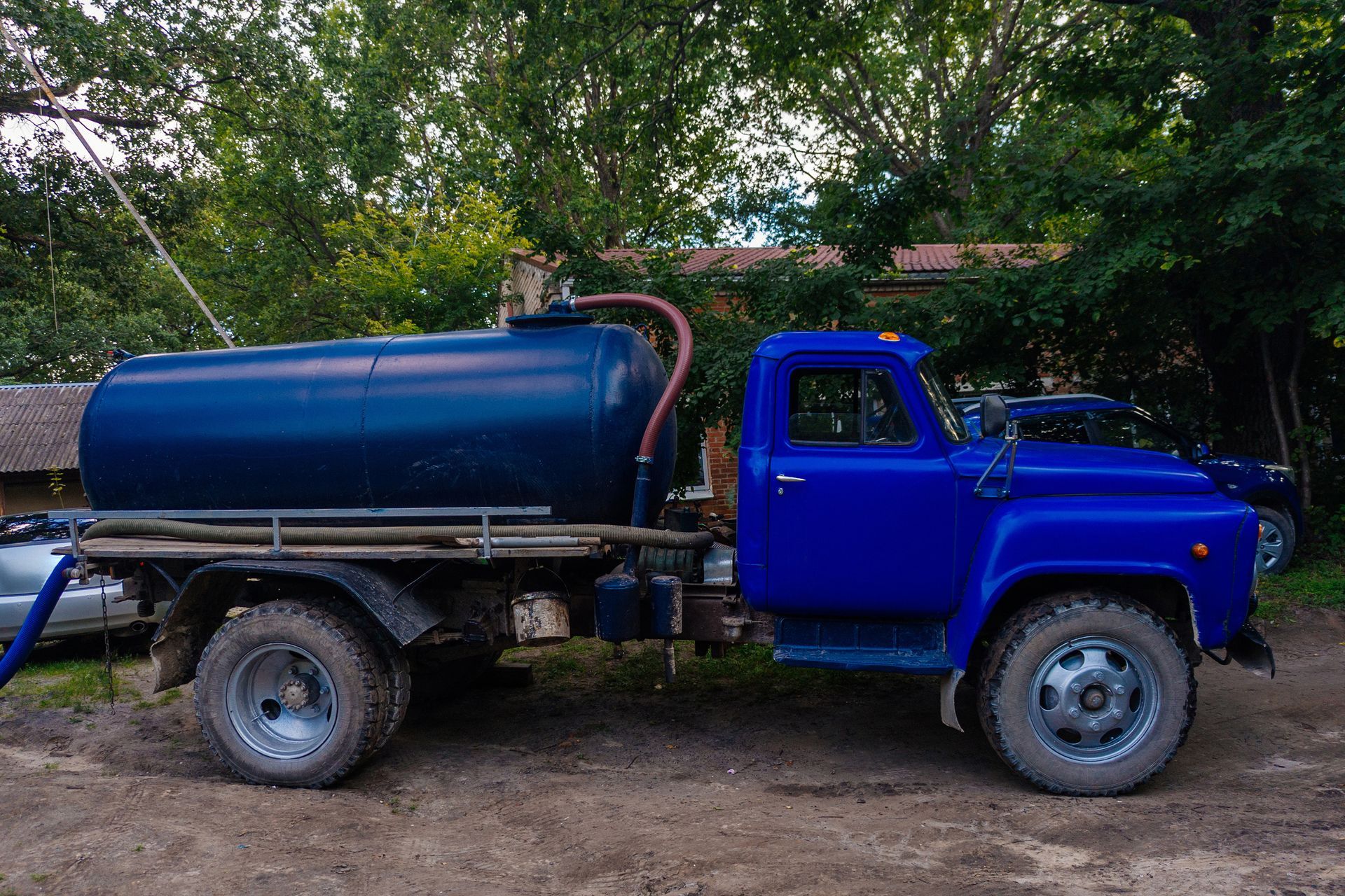 Blue vintage pickup truck with a large cylindrical tank parked on a dirt path beside trees