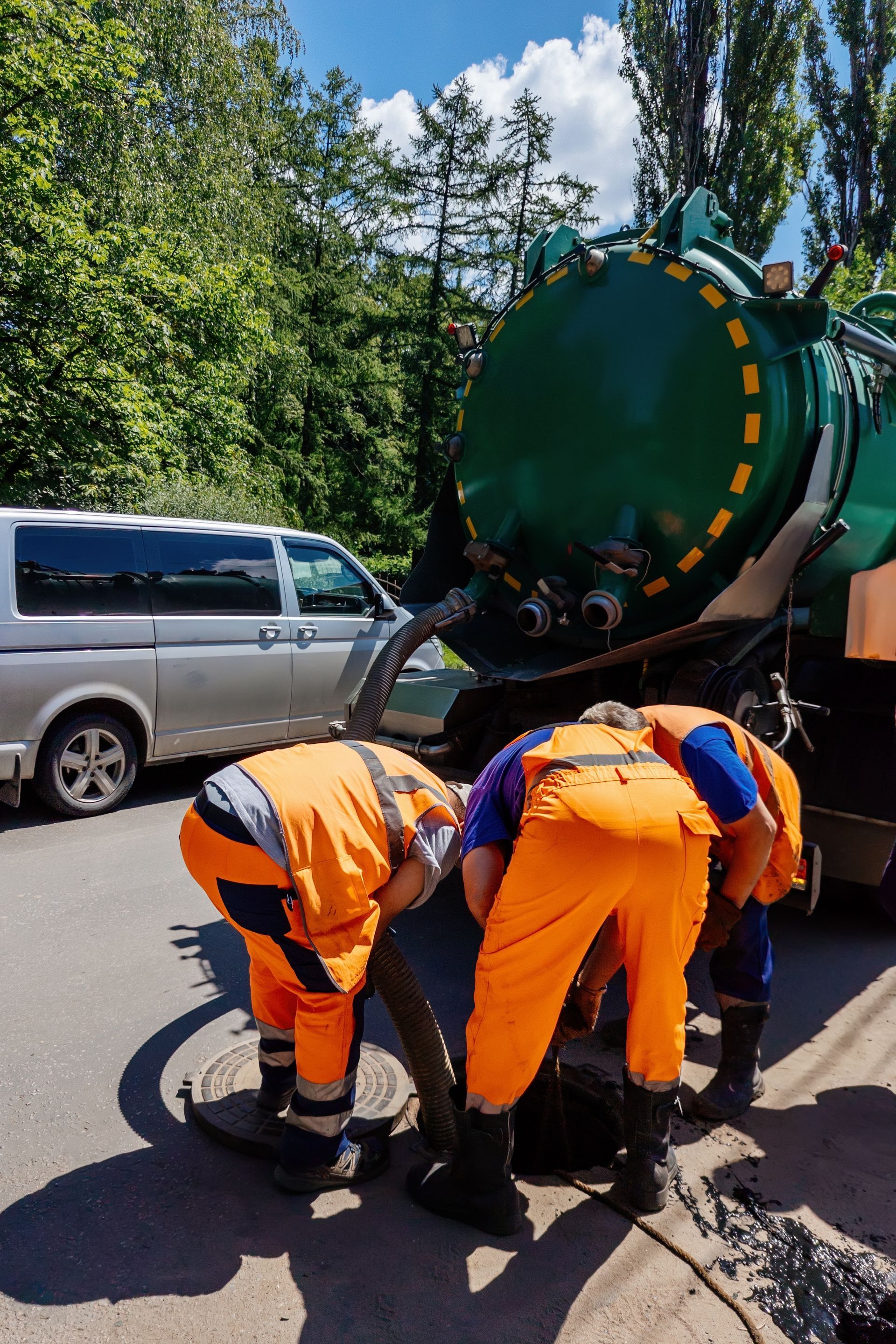 Workers in orange uniforms repair a green utility truck on a roadside in daylight.
