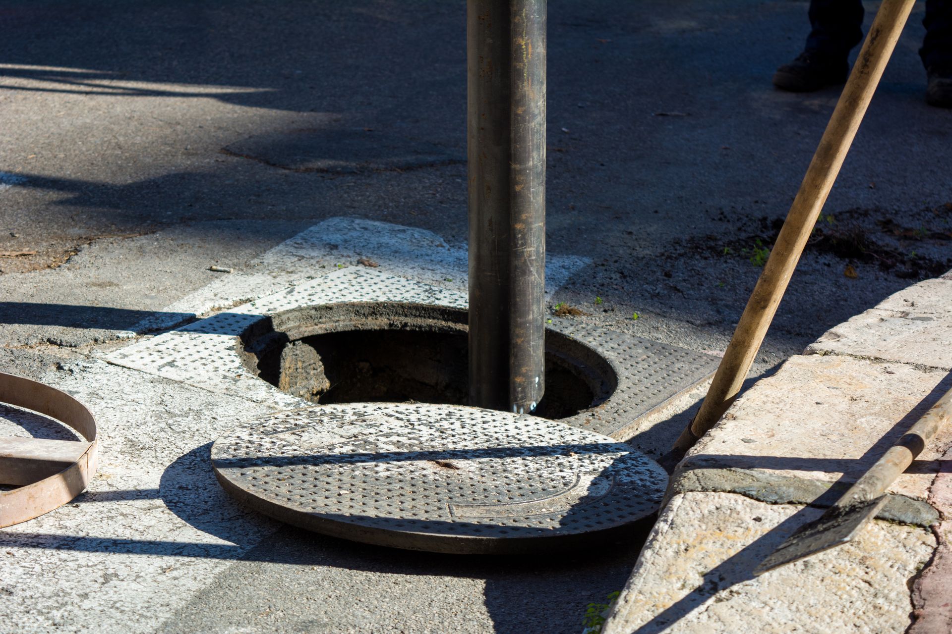 Open manhole in cracked sidewalk with a metal cover propped nearby and a pole in the hole