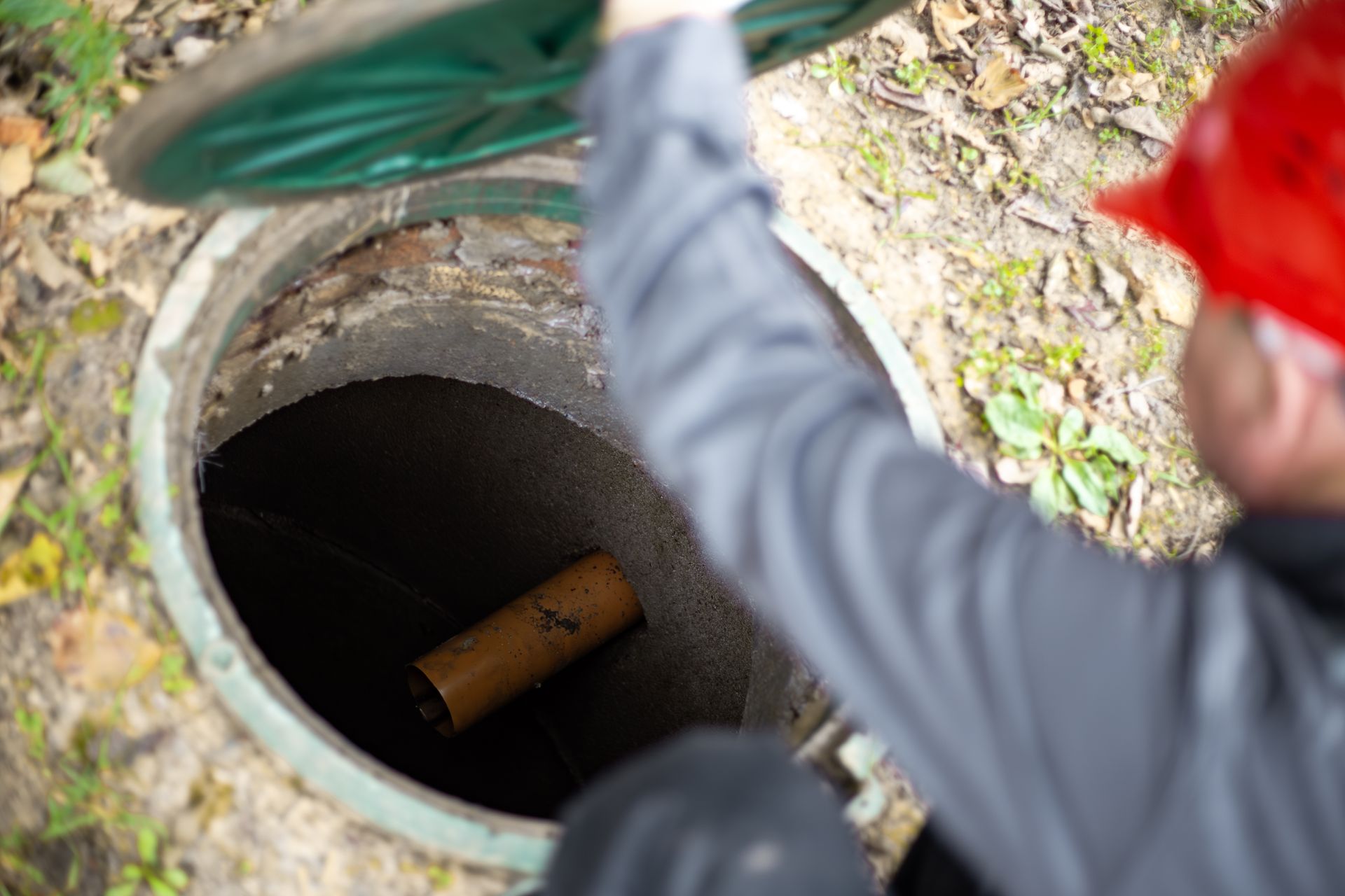 Person looking into open manhole with a green tool and red helmet nearby