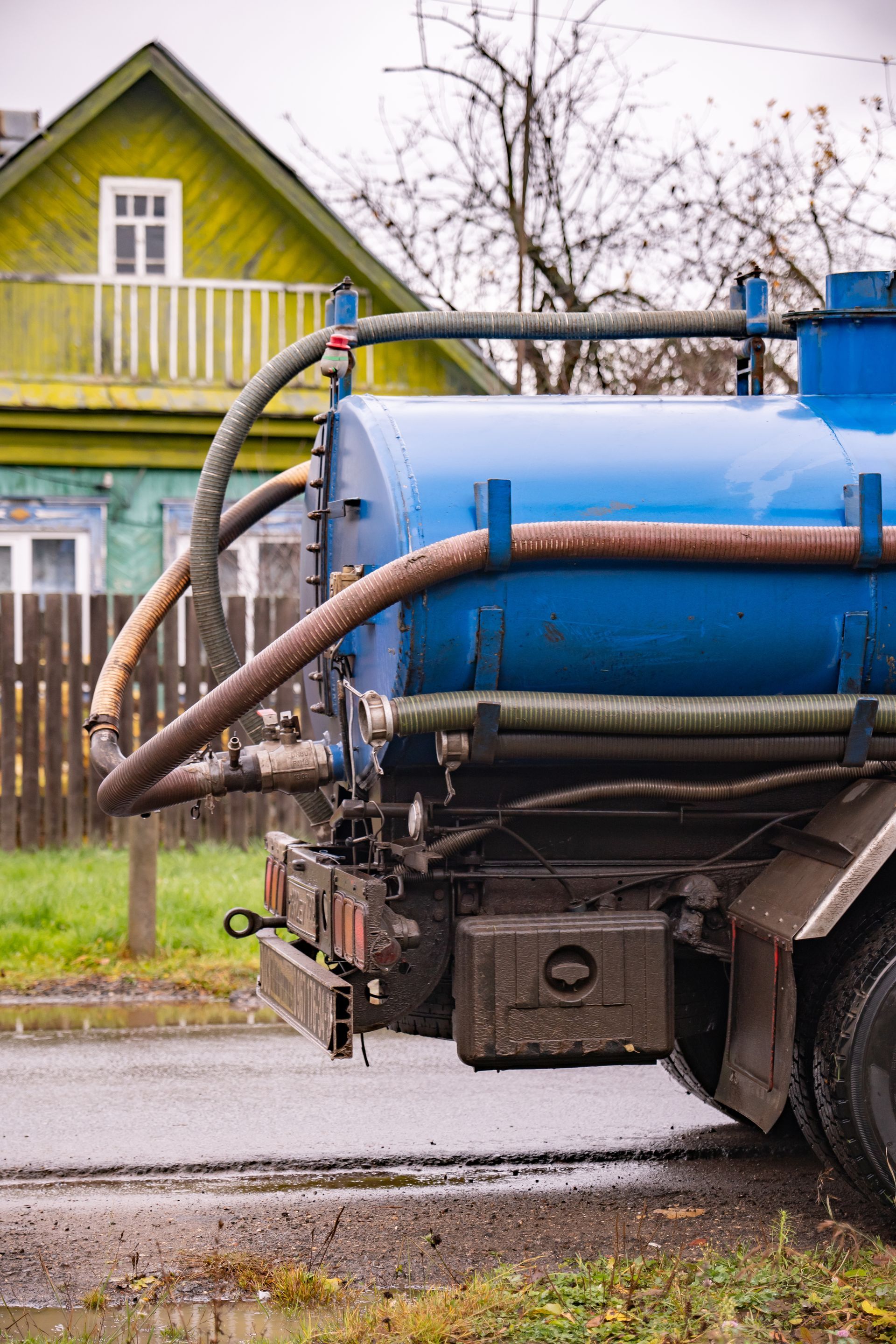 Blue vacuum truck parked by a yellow house on a wet street, with hoses and tanks visible