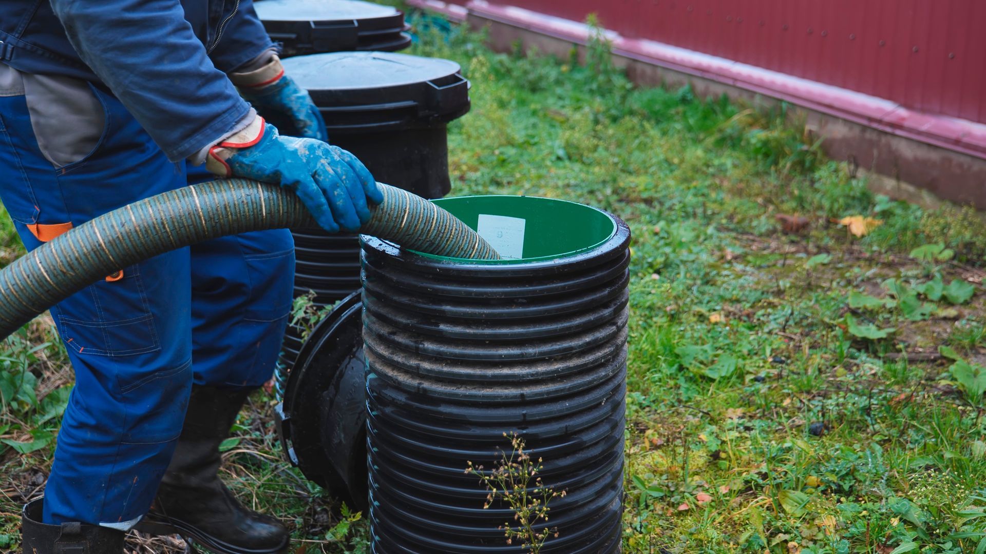 Worker in blue gloves pouring liquid into a black corrugated barrel outdoors on grass