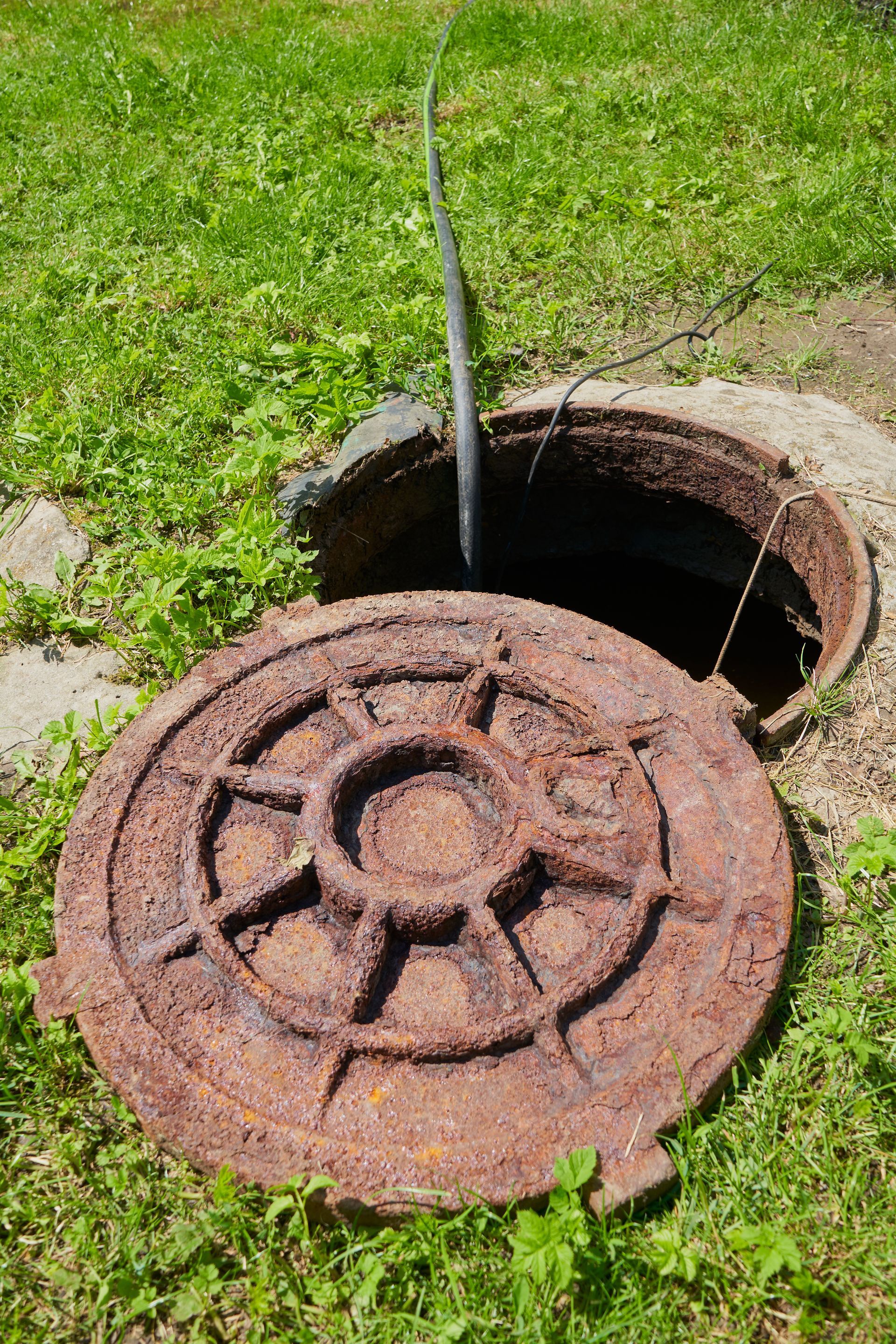 Rusty manhole cover open on grass beside a dark sewer opening