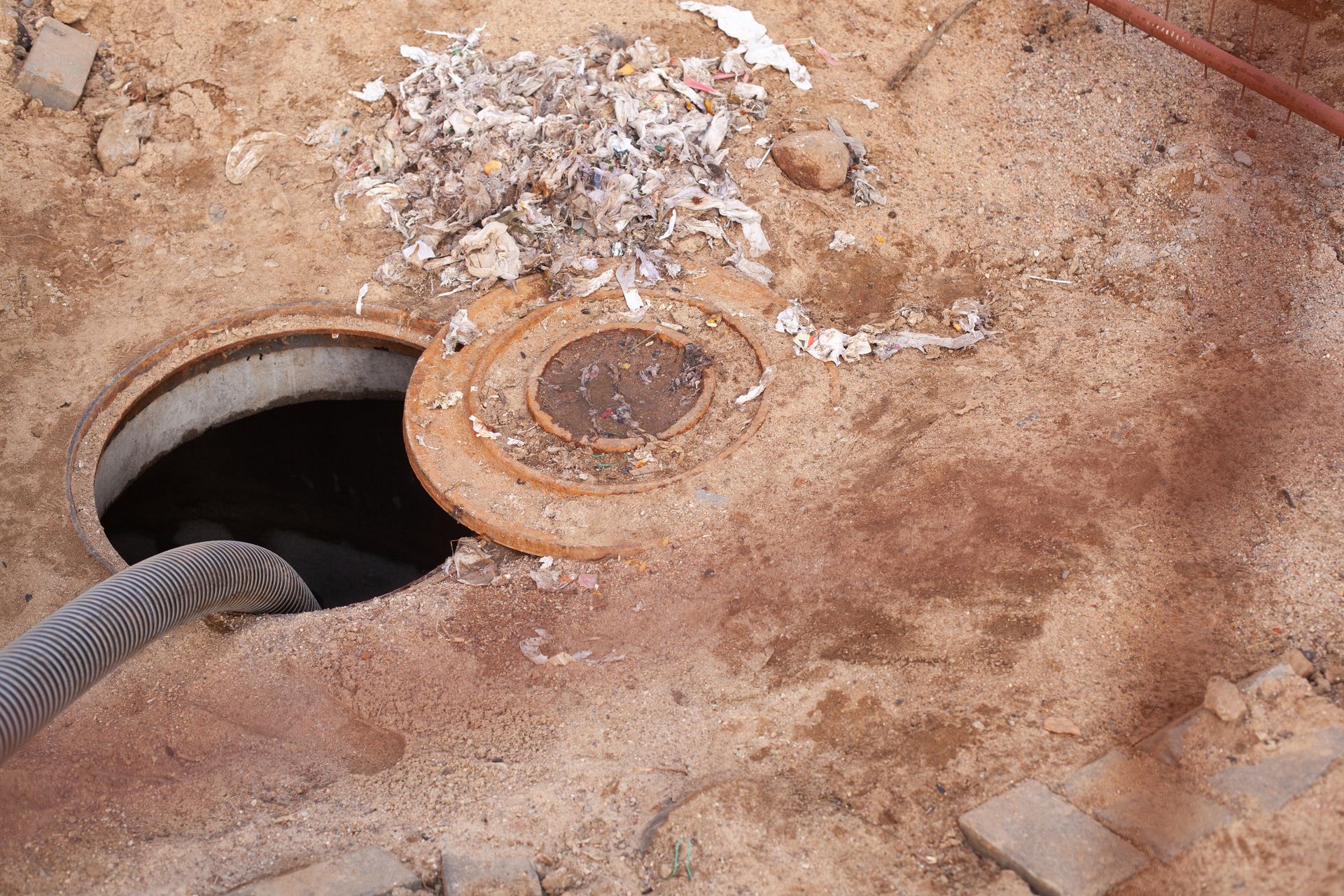 Open manhole in dirt with a black hose and debris around the cover